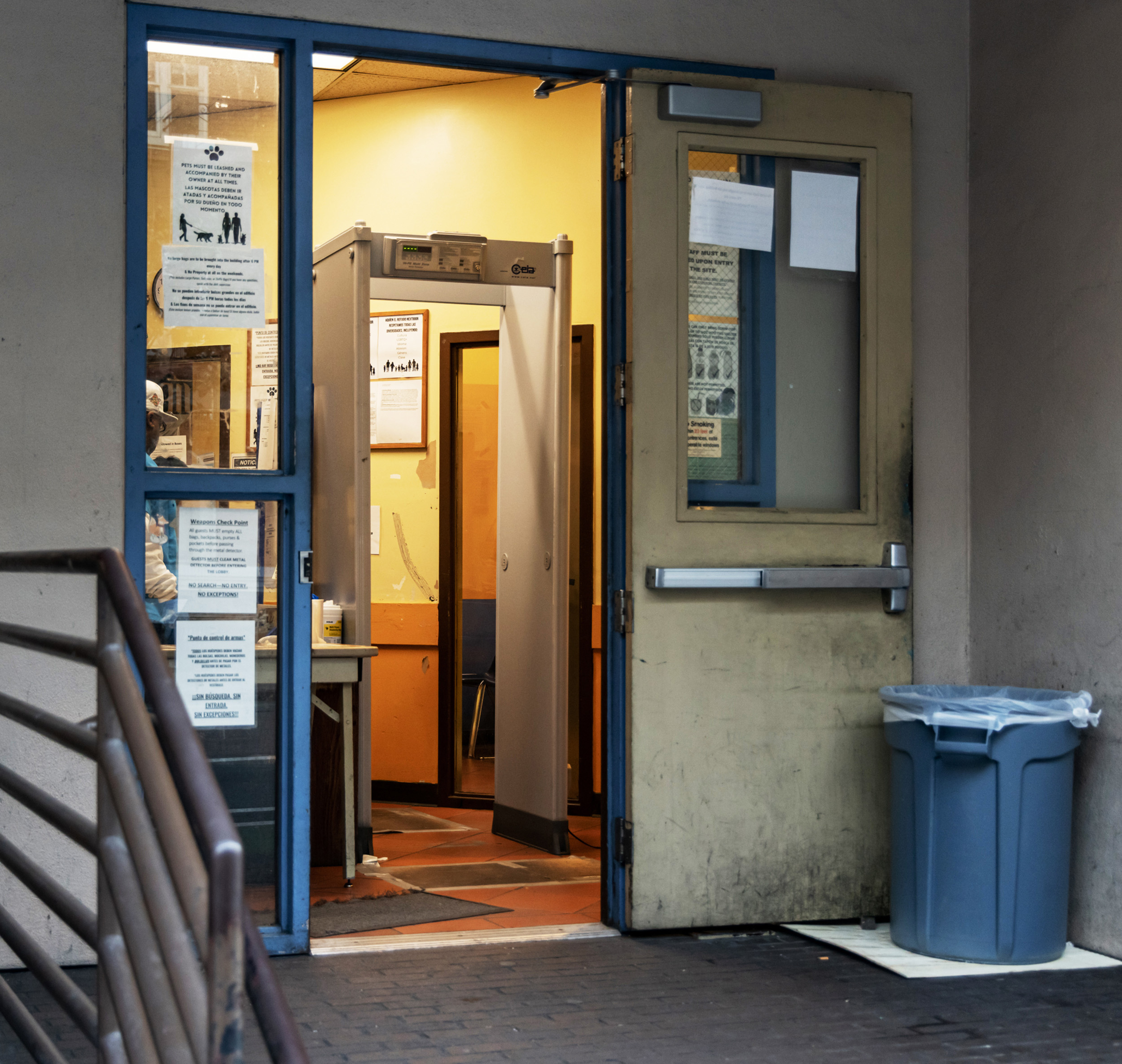 The image shows an open doorway with a metal detector inside. Posters are on the glass windows, and a trash can is outside. The room appears to be an entryway or lobby.