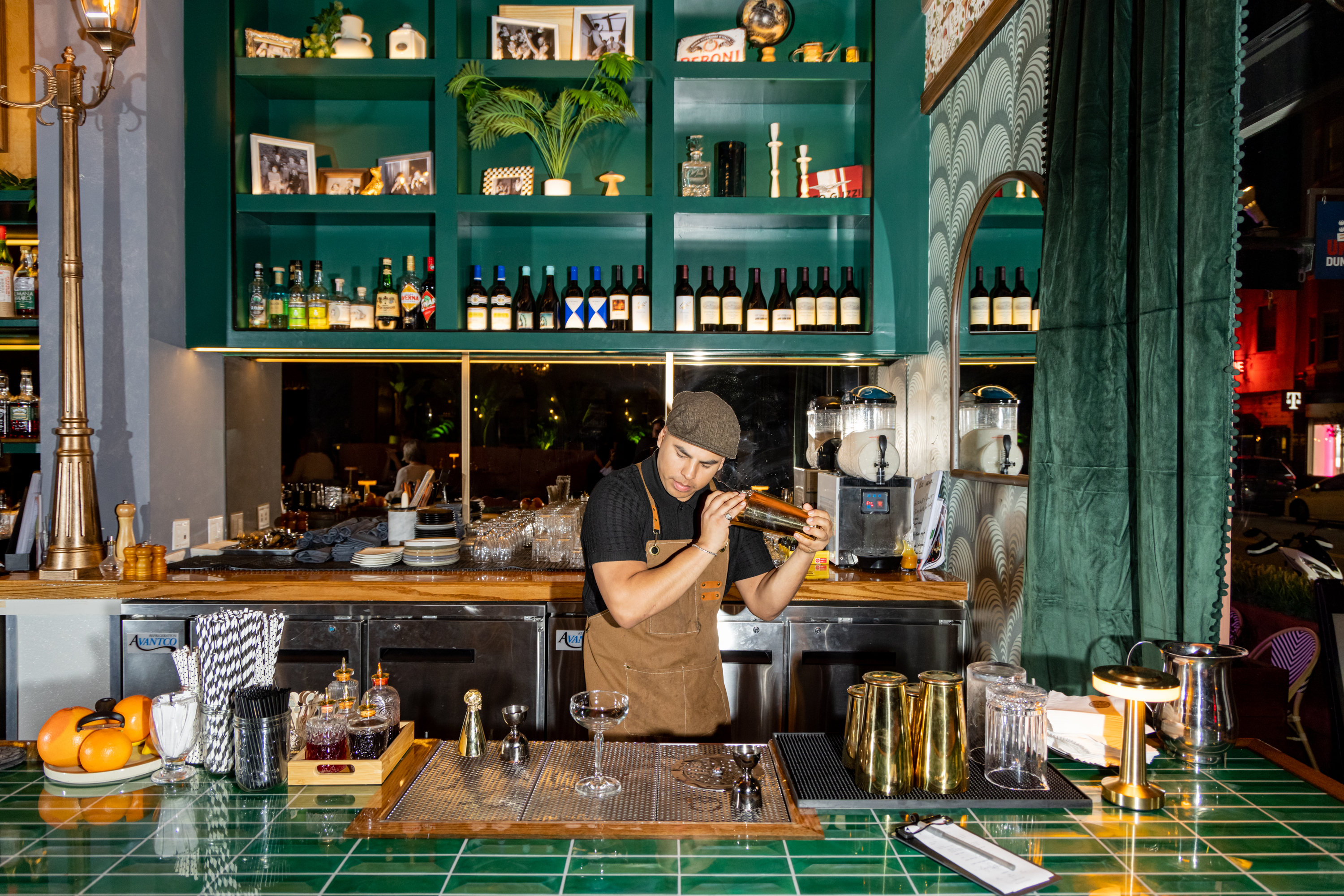 A bartender in a flat cap mixes a cocktail at a green-tiled bar with shelves of bottles and decorative items. The bar is well-lit and neatly arranged.