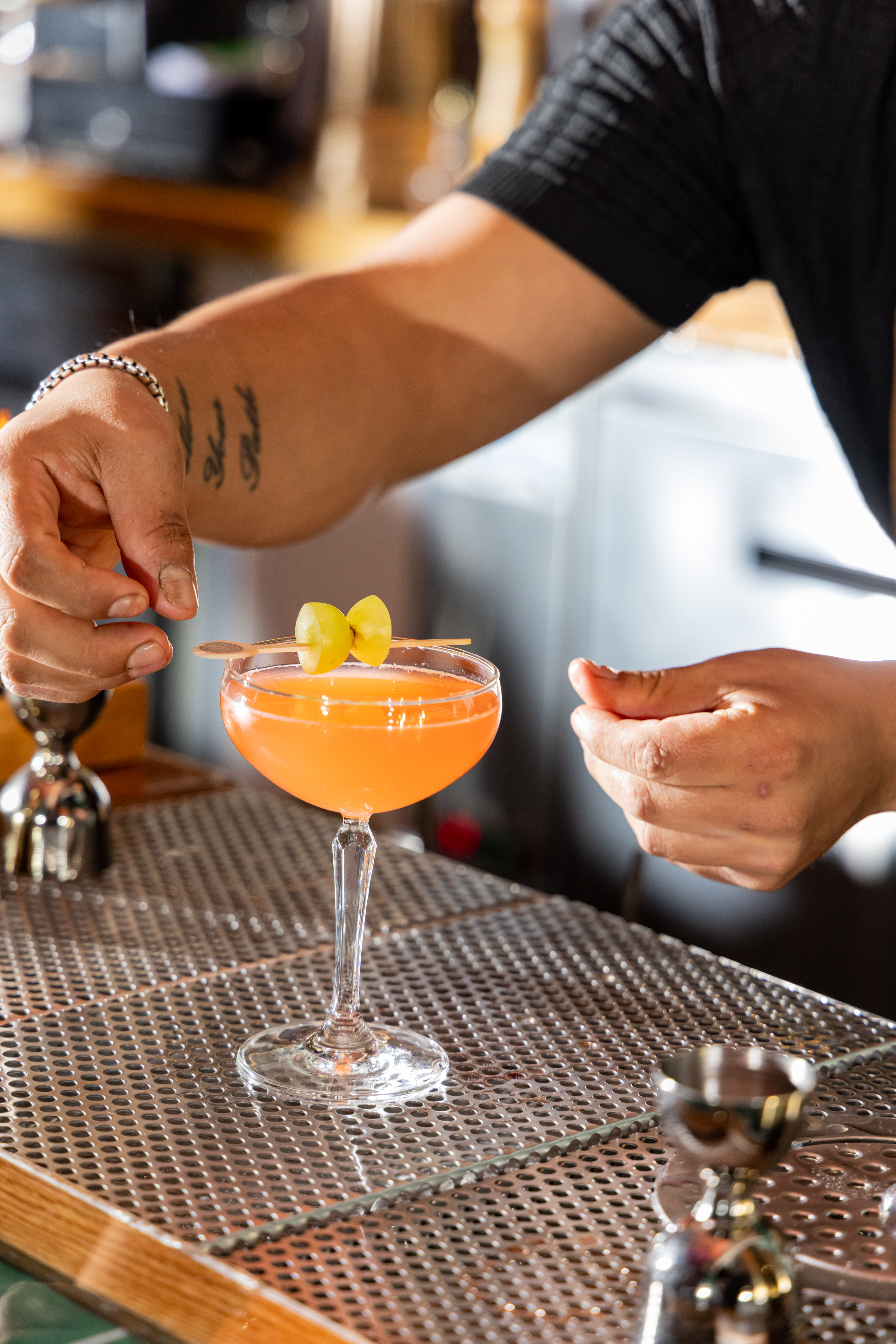 A person garnishes an orange cocktail in a coupe glass with a toothpick holding small green fruits. The scene is set on a metal bar counter.