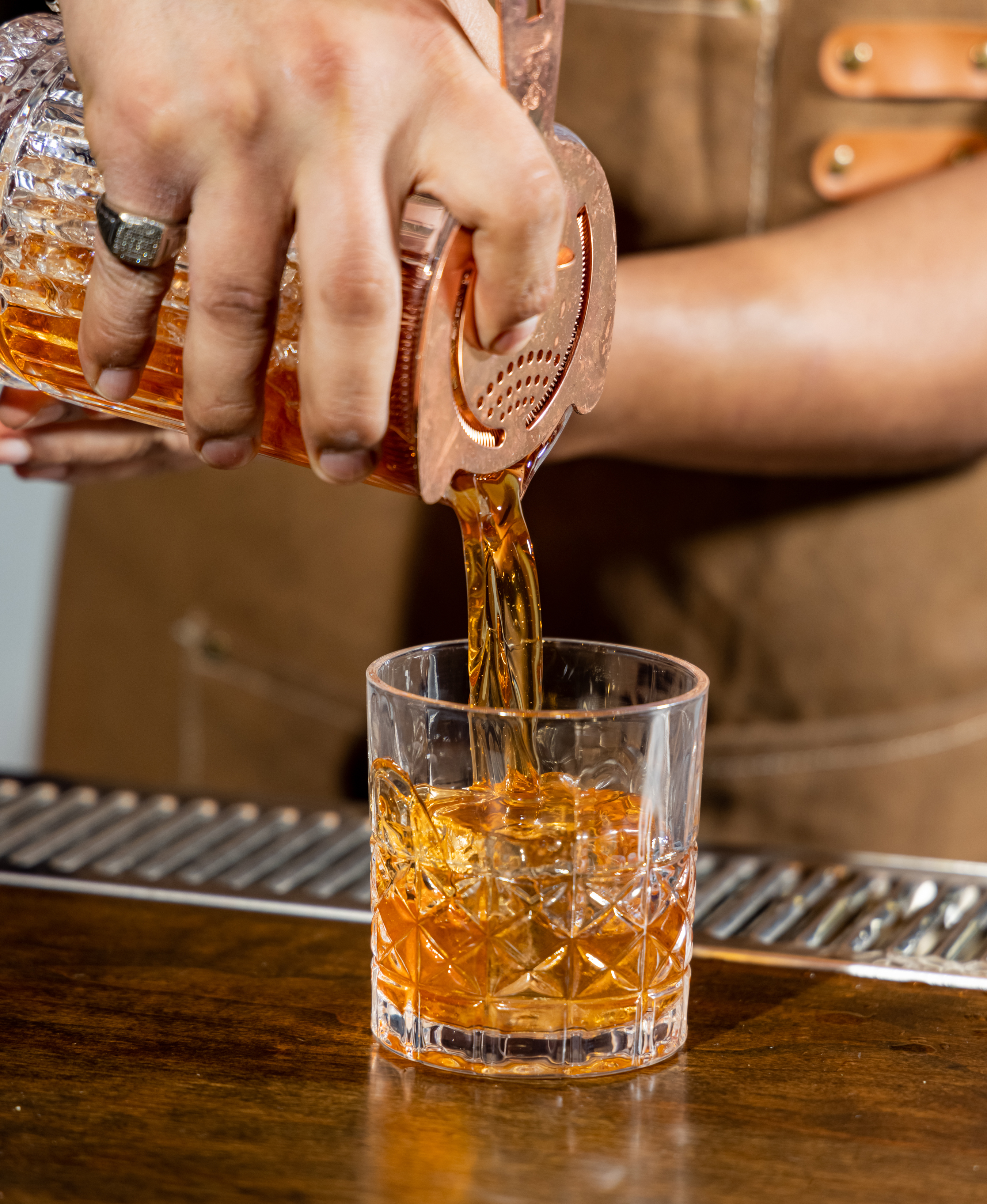 A person is pouring a vibrant amber liquid into a textured glass using a strainer. The background shows a bar counter and a person with a brown apron.