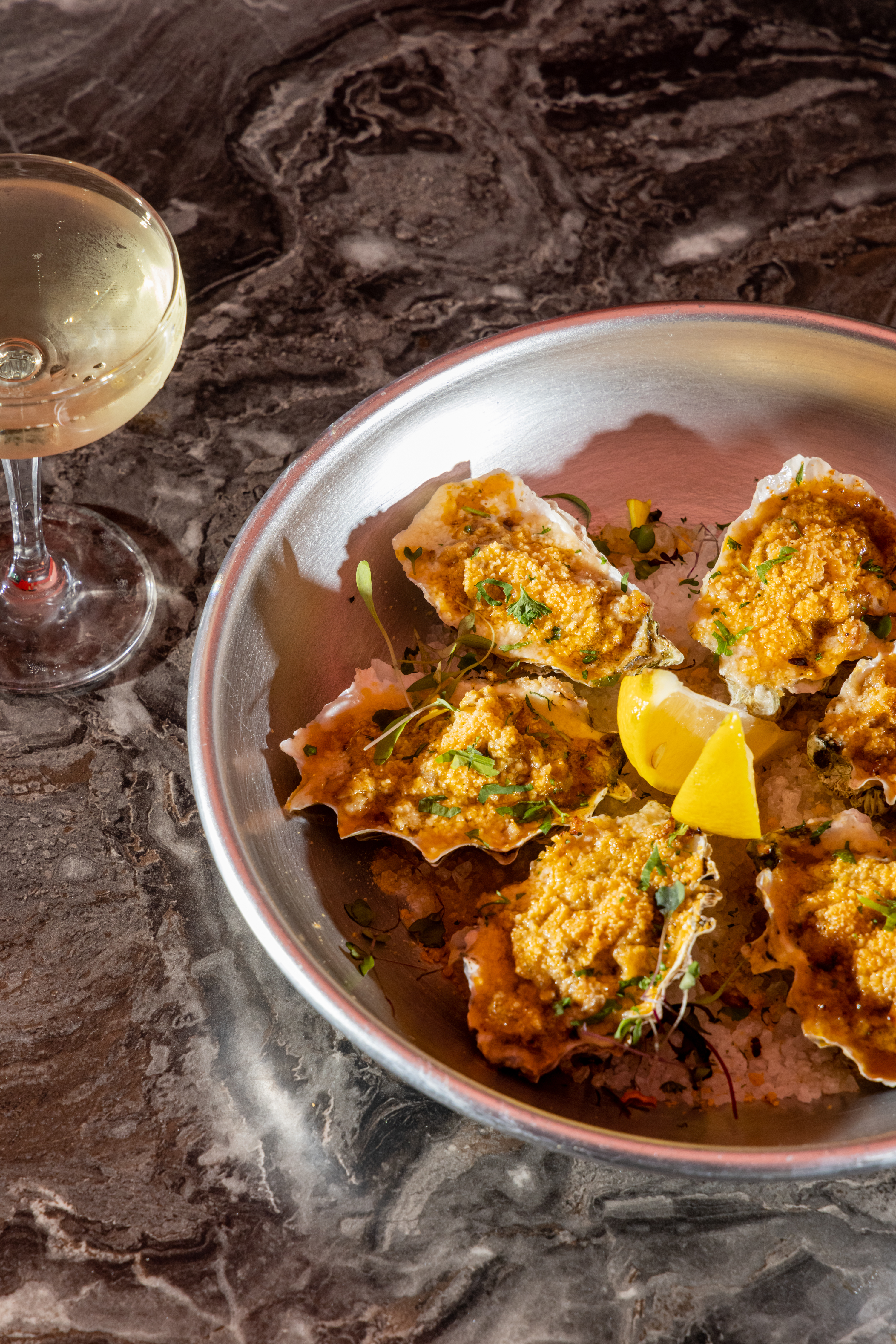 A metal dish holds baked oysters topped with a golden crust, garnished with herbs and lemon slices. Beside it is a glass of white wine on a marble surface.