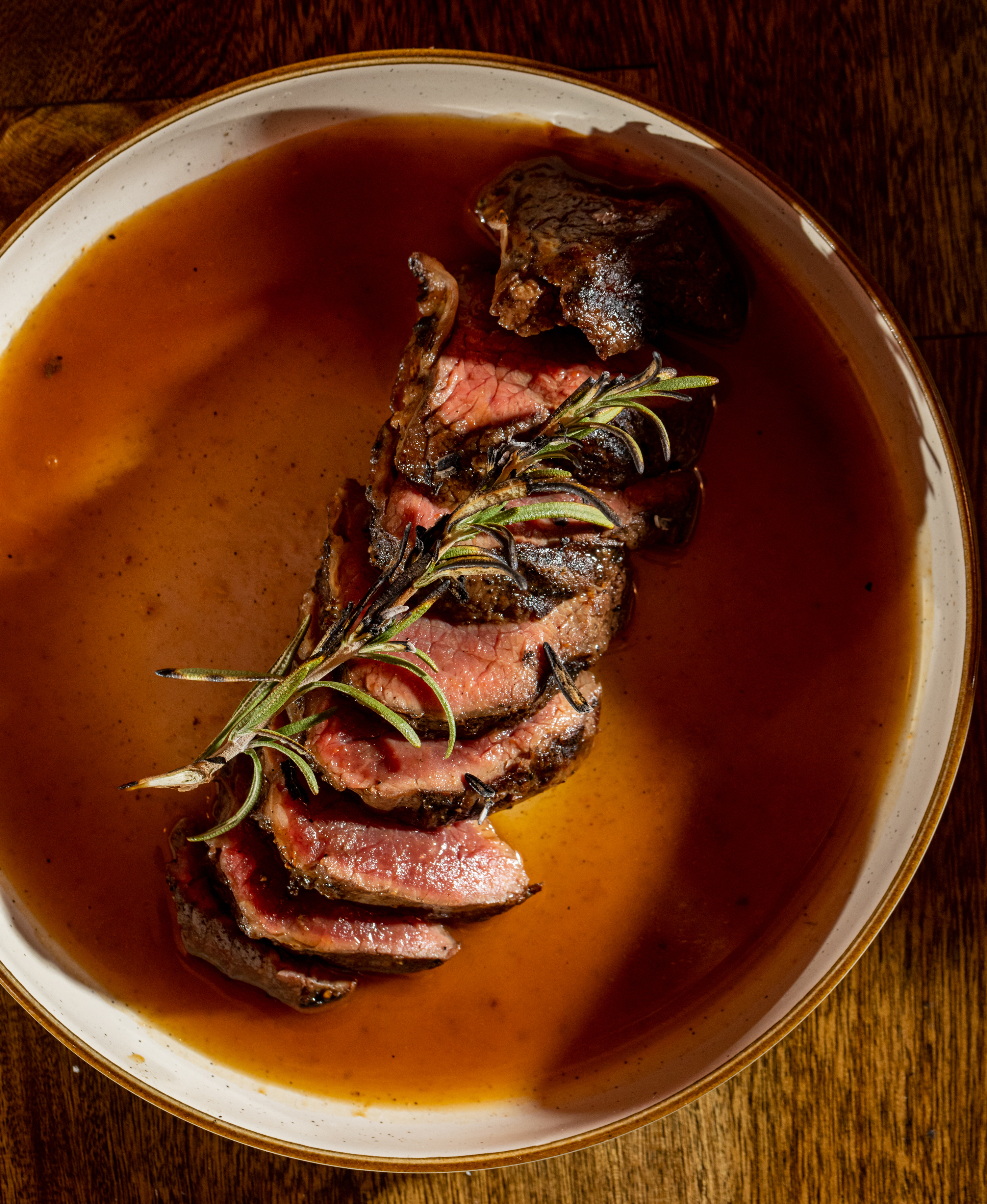 Slices of grilled steak, cooked rare, are arranged in a shallow bowl filled with a rich brown sauce. A sprig of rosemary rests on top.