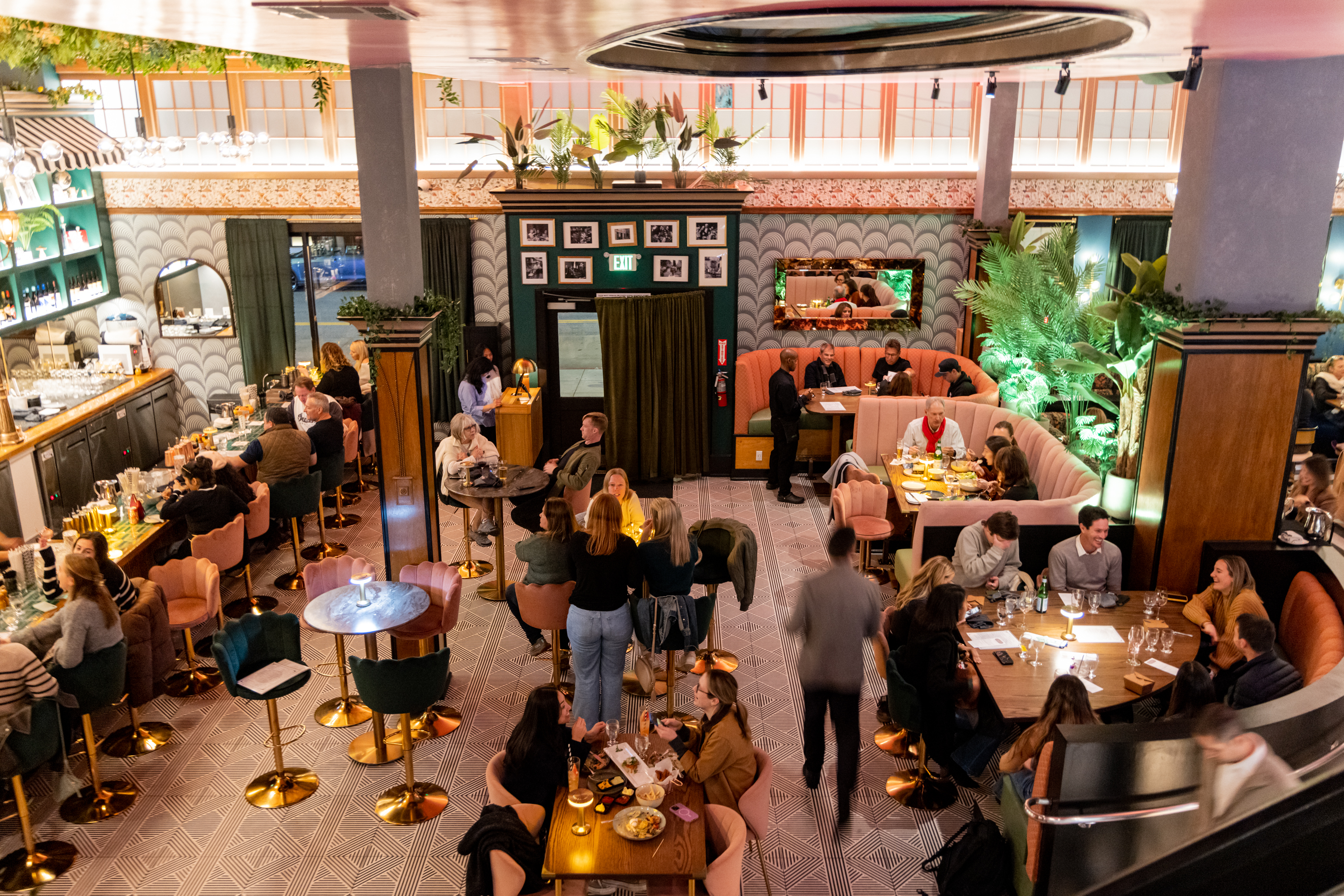 The image shows a bustling, stylish restaurant interior with people dining at various tables and a bar. The space is decorated with plants and ambient lighting.