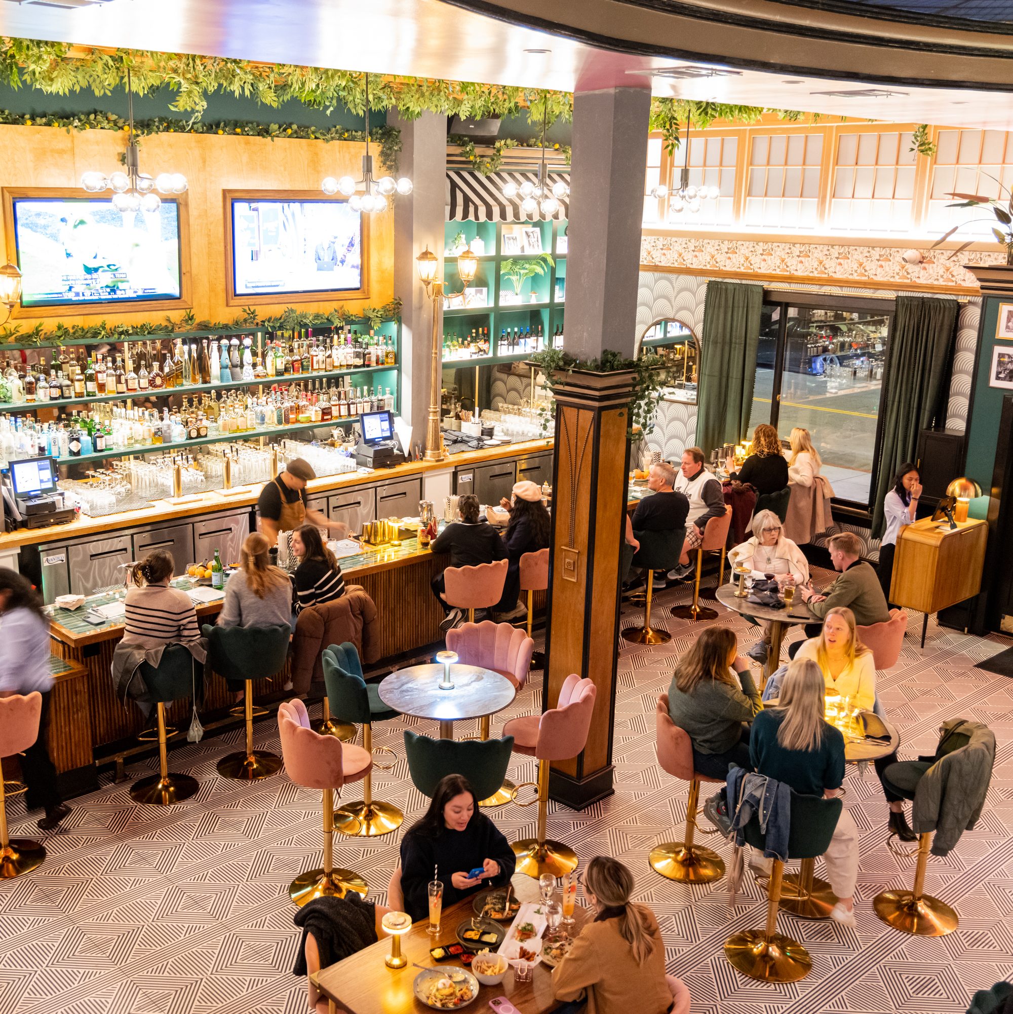 The image shows a lively, stylish bar with people seated at tables and the bar counter. The space is decorated with green plants and warm lighting.