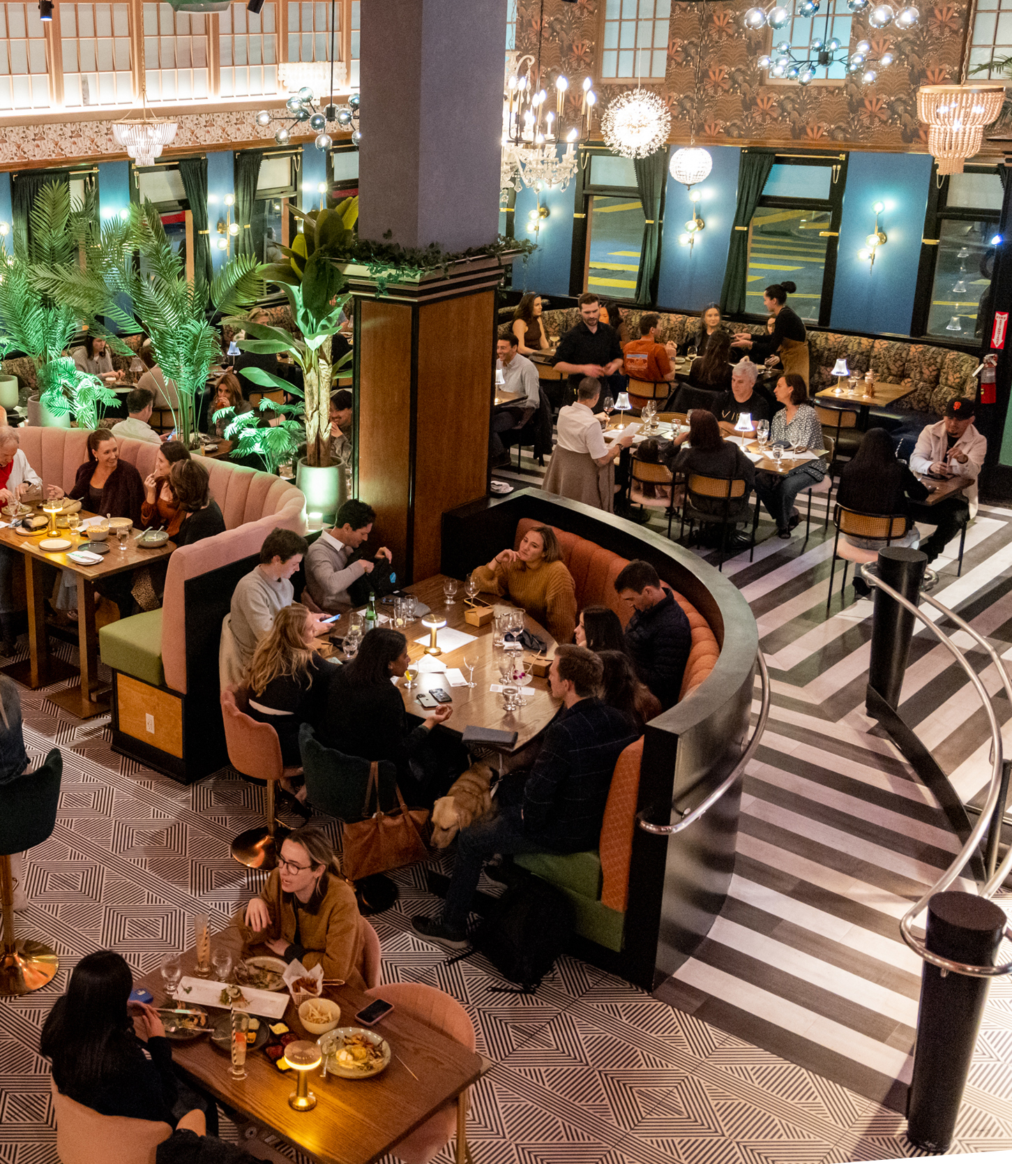 A lively restaurant scene shows people dining at various tables, surrounded by lush plants, elegant chandeliers, and patterned floors in a stylish interior.