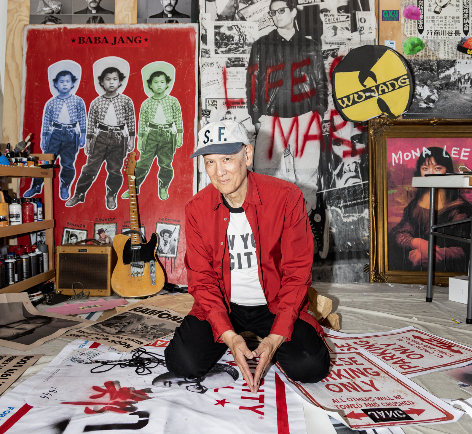 A person wearing a red jacket and &quot;S.F.&quot; cap sits on the floor in an art-filled room. The space has posters, a guitar, and colorful graffiti.