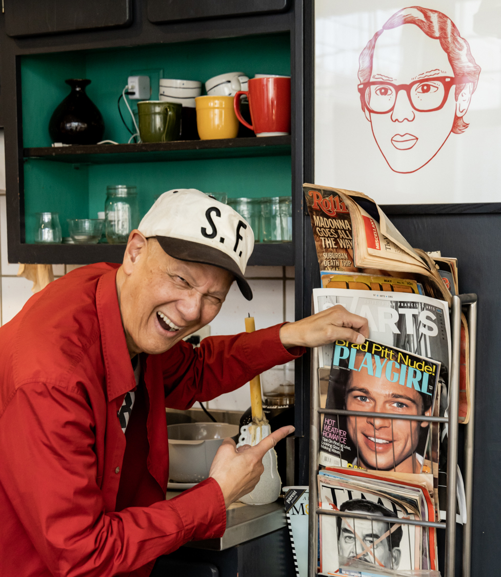 A smiling person in a white cap and red shirt points at a magazine rack with various covers. Colorful mugs are on shelves, and a framed drawing is on the wall.