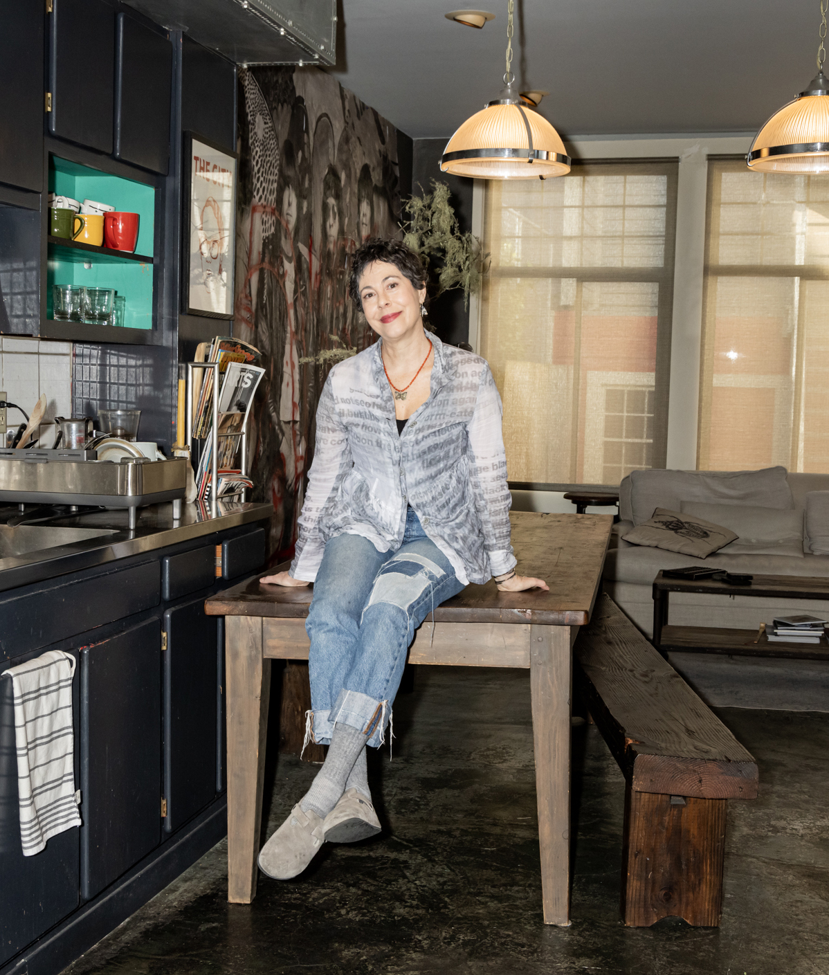 A person sits casually on a wooden table in a cozy kitchen with dark cabinets, colorful mugs, and artistic wallpaper. Warm lighting and large windows are in the background.