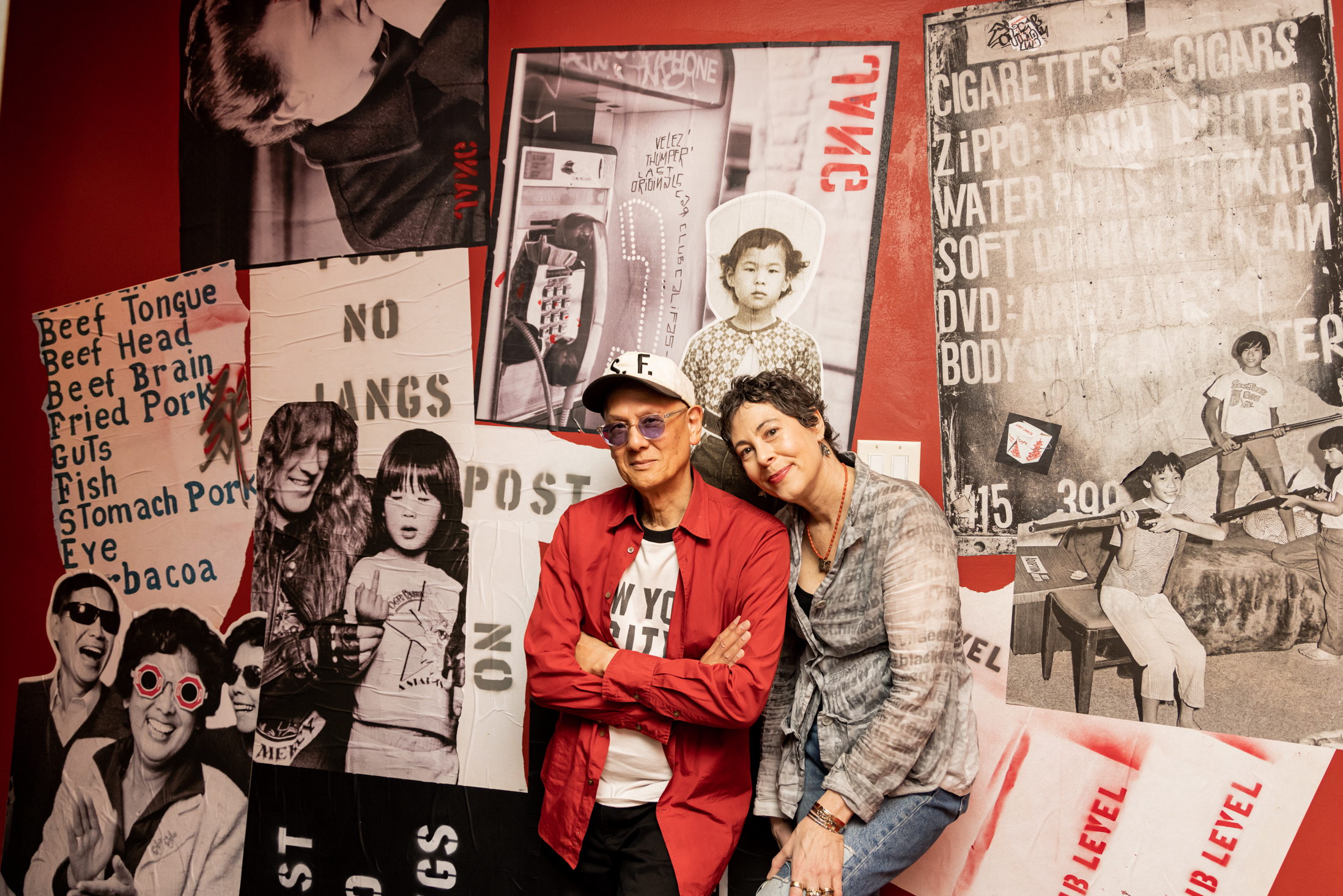 A man in a red shirt and hat stands with a woman leaning on him, surrounded by large black-and-white posters on a red wall, featuring various images and texts.