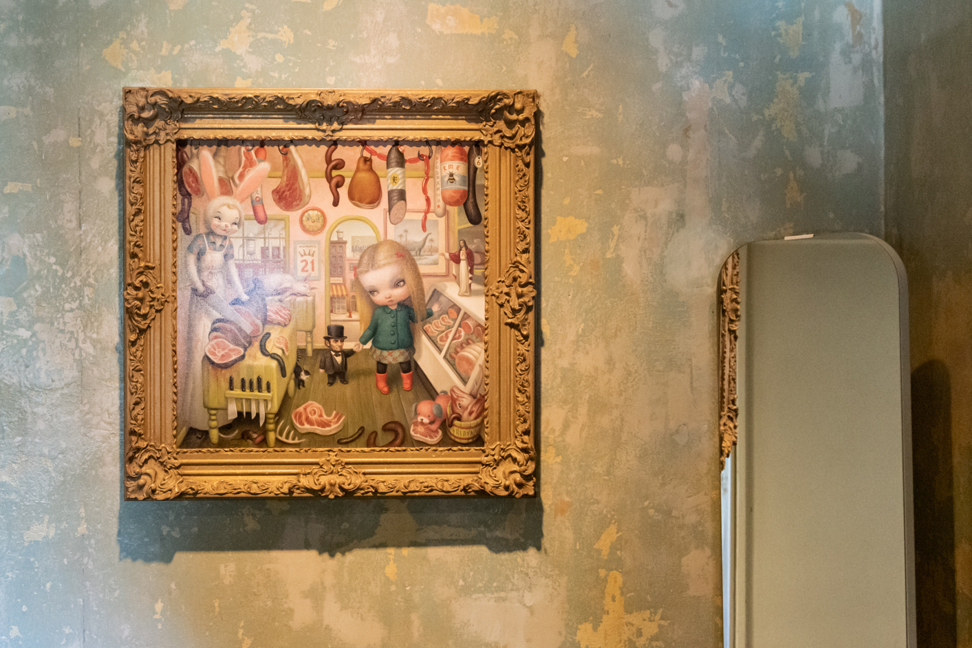 A framed painting on a textured wall depicts a whimsical scene with doll-like figures in a butcher shop. There's a mirror reflecting part of the wall.