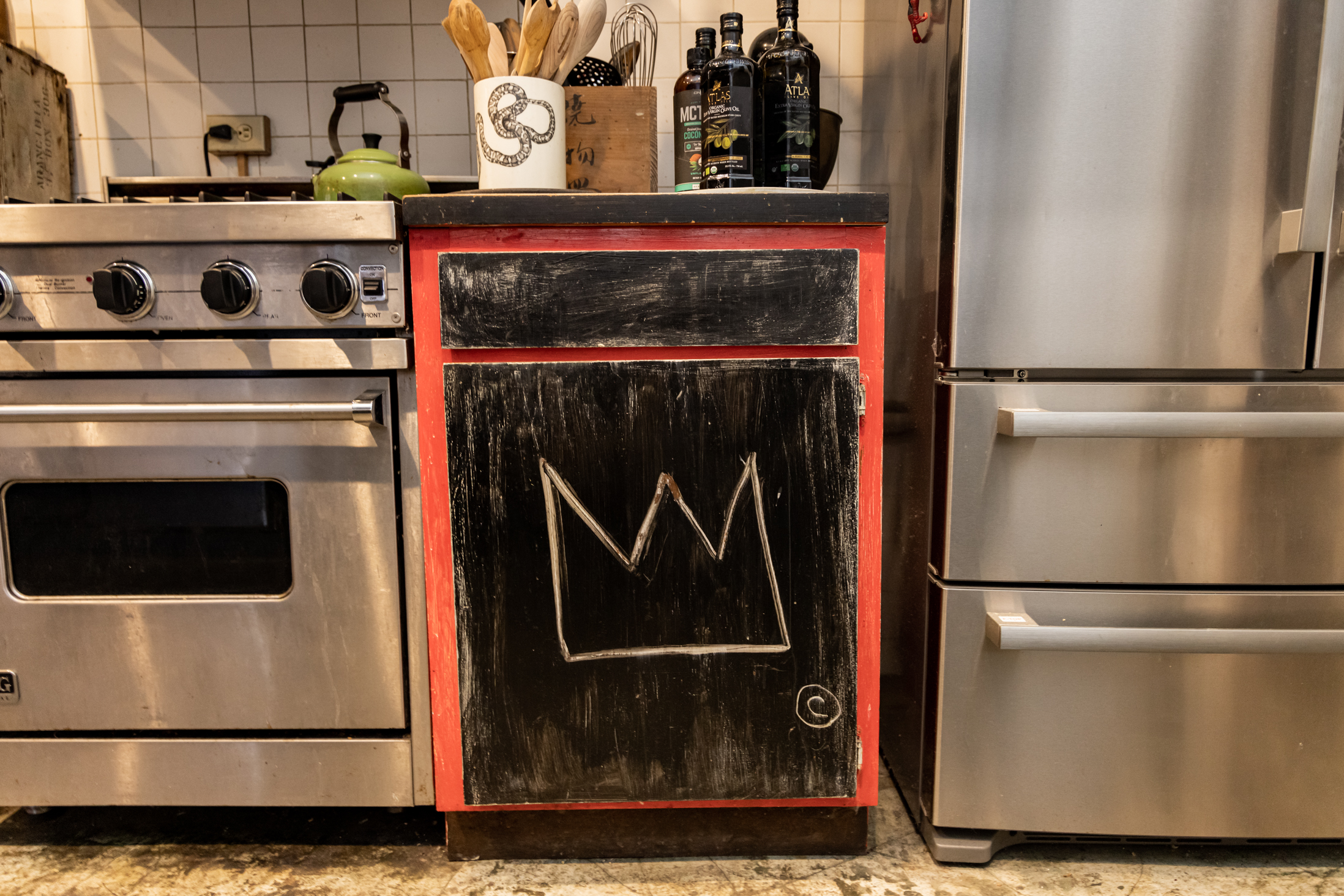 The image shows a modern kitchen with a stainless steel oven, a fridge, and a cabinet with a black chalkboard front featuring a drawn crown. Various kitchen items are on the counter.