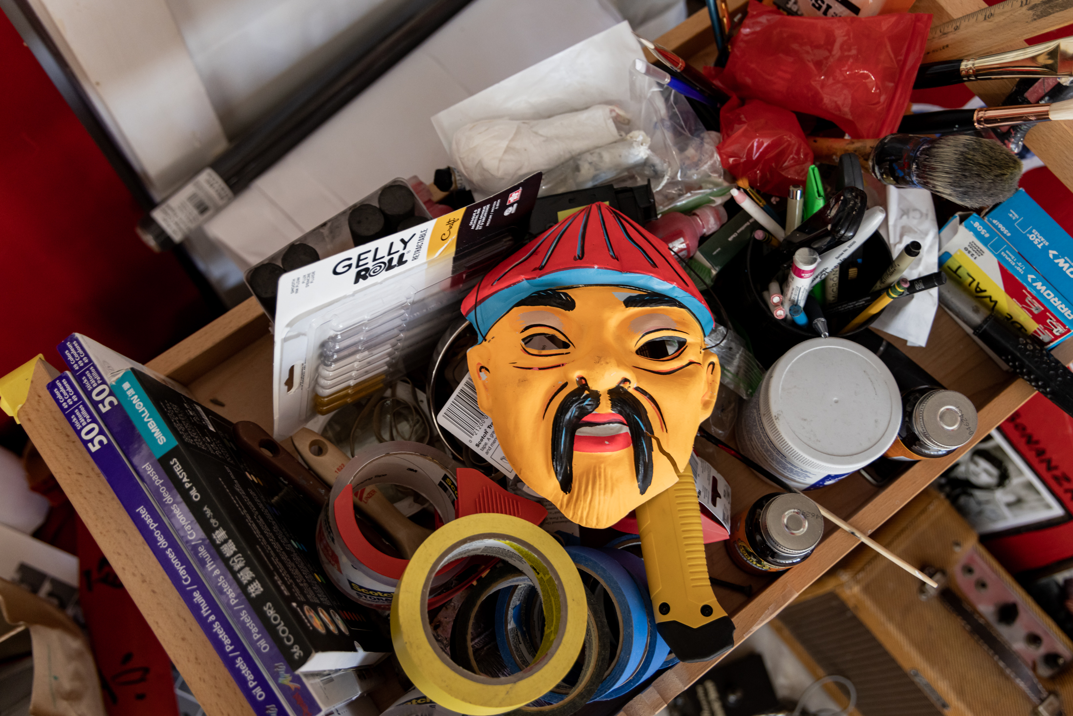 A cluttered drawer is filled with art supplies, tapes, pens, and paint, topped with a colorful, stylized mask featuring a mustache and hat.