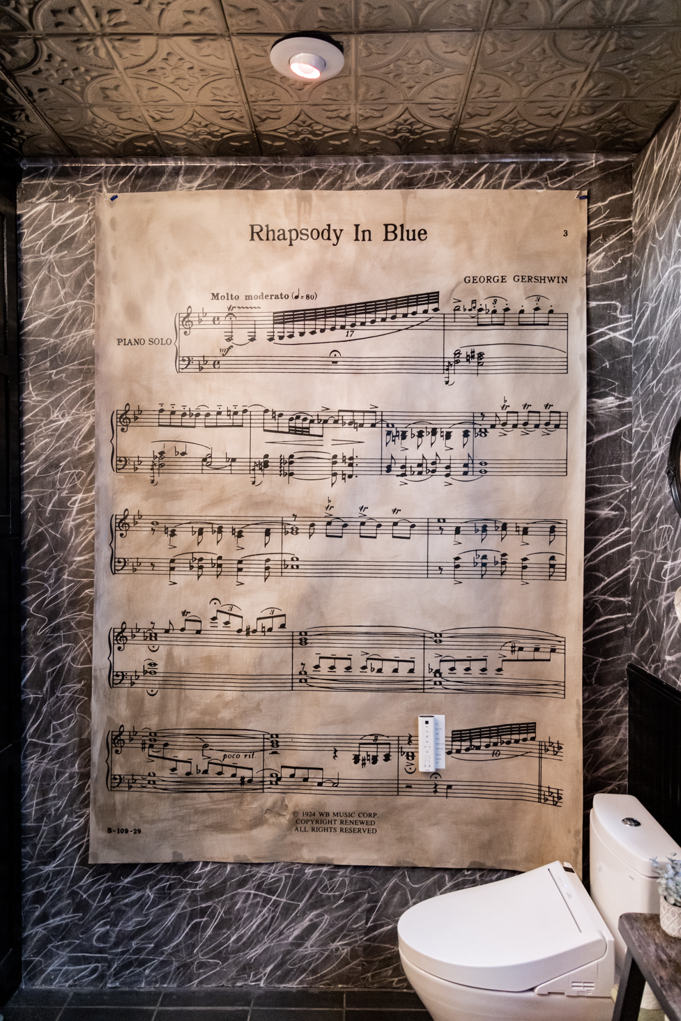 A large sheet of &quot;Rhapsody In Blue&quot; music by George Gershwin is displayed on a textured wall above a modern toilet in an ornately designed restroom.