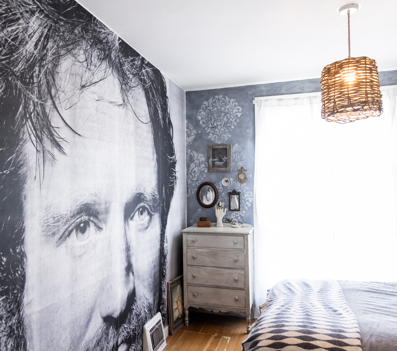 The image shows a bedroom with a large grayscale mural of a man's face on the wall. There's a dresser with mirrors, decorative items, and a woven hanging light.