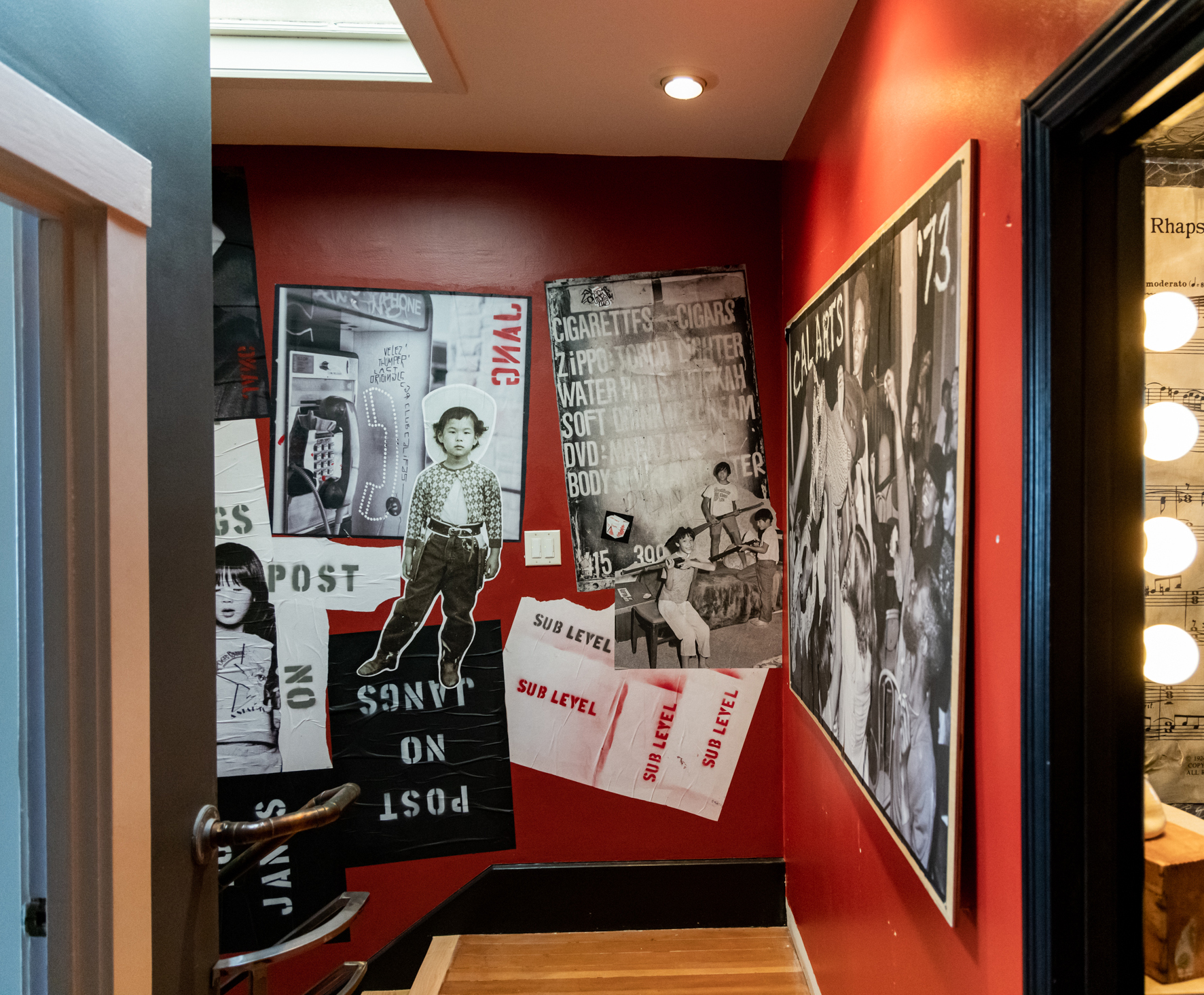 The image depicts a hallway with red walls featuring various collaged posters, including images of people and text. The floor is wooden, and there's a lit-up area.