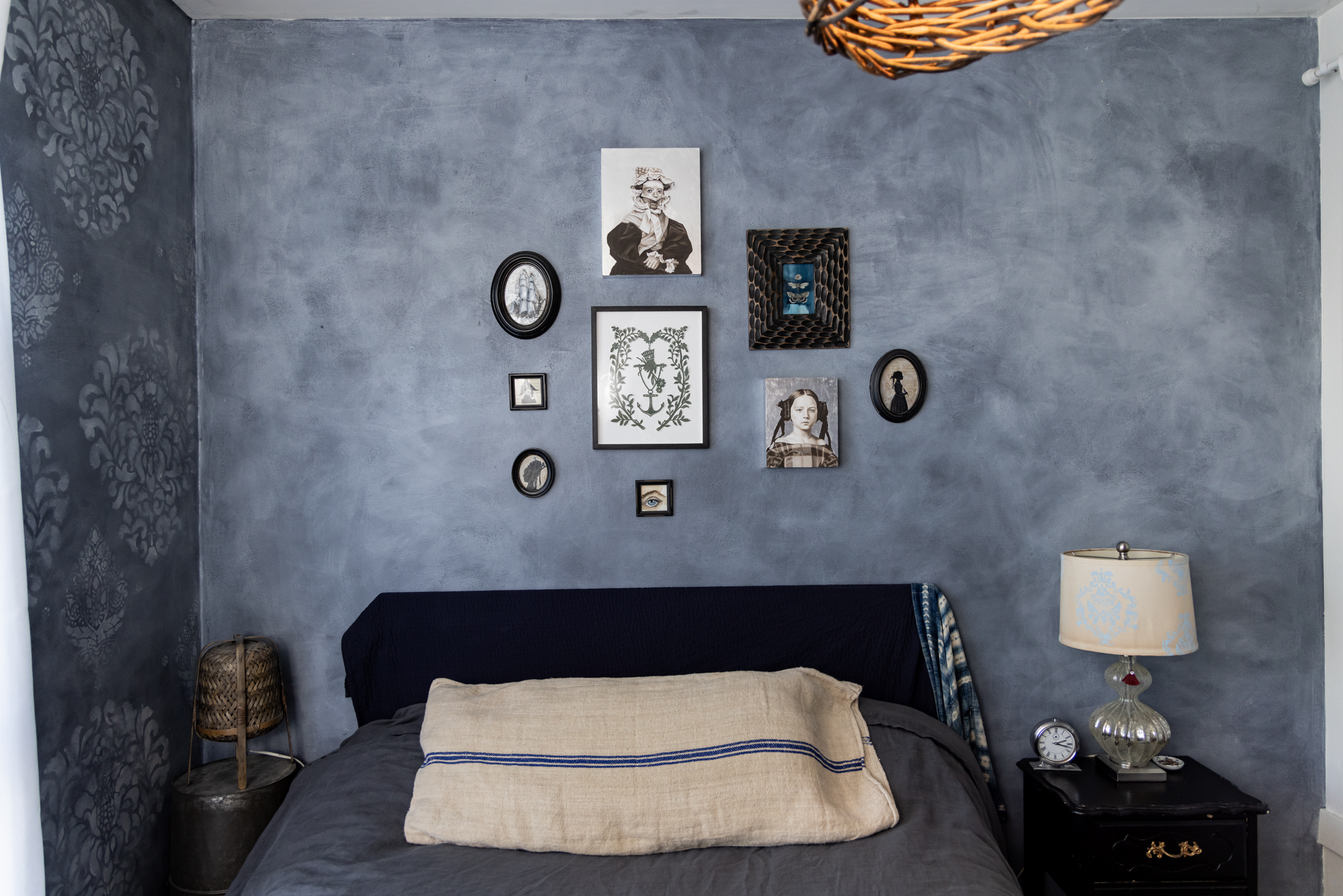 A bedroom features a navy bed with beige pillows. The wall behind displays various framed art pieces. A lamp and clock sit on a nightstand, and a wicker light hangs above.