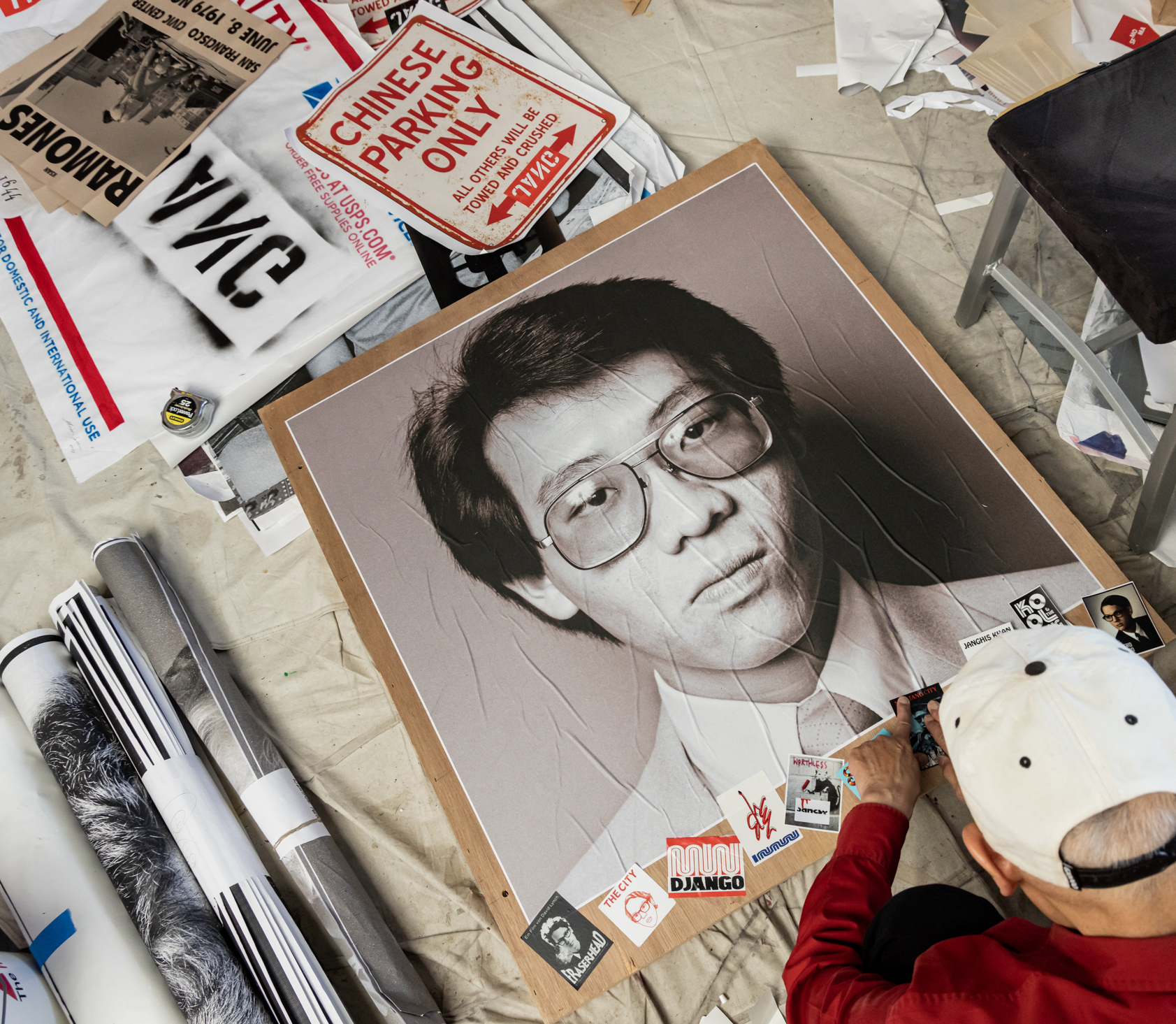 A person in a red shirt and white cap works on a large black-and-white portrait on the floor, surrounded by posters and papers, including a &quot;Chinese Parking Only&quot; sign.