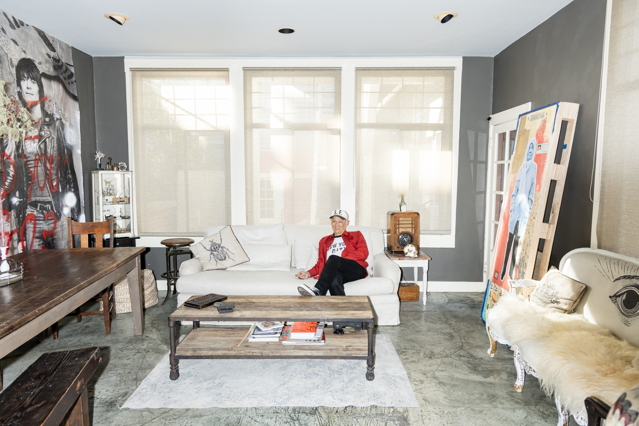 A person in a red jacket sits on a white couch in a room with large windows, gray walls, and eclectic decor, including a mural and a vintage radio.