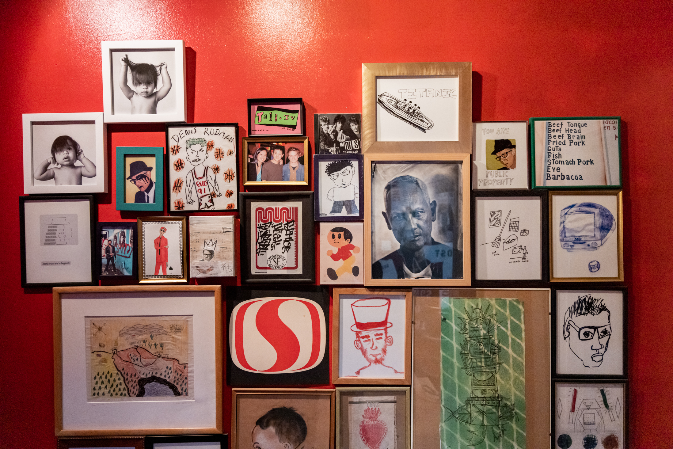 A vibrant red wall features a collage of diverse framed artwork, including drawings, paintings, and photos, showcasing abstract, realistic, and playful styles.