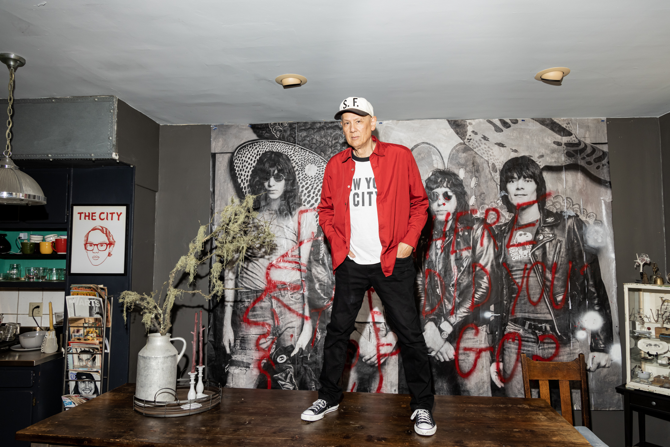 A person in a red jacket and cap stands on a wooden table in front of a mural of a rock band. The room has a retro vibe with kitchen items around.