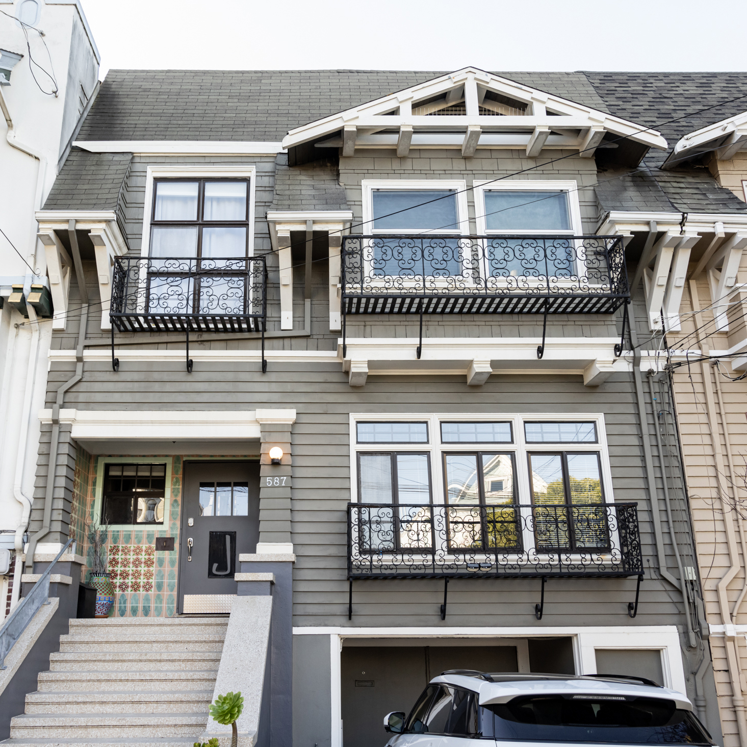 A two-story gray house features decorative black railings, two balconies, and a welcoming entrance with stairs. A car is parked in front.