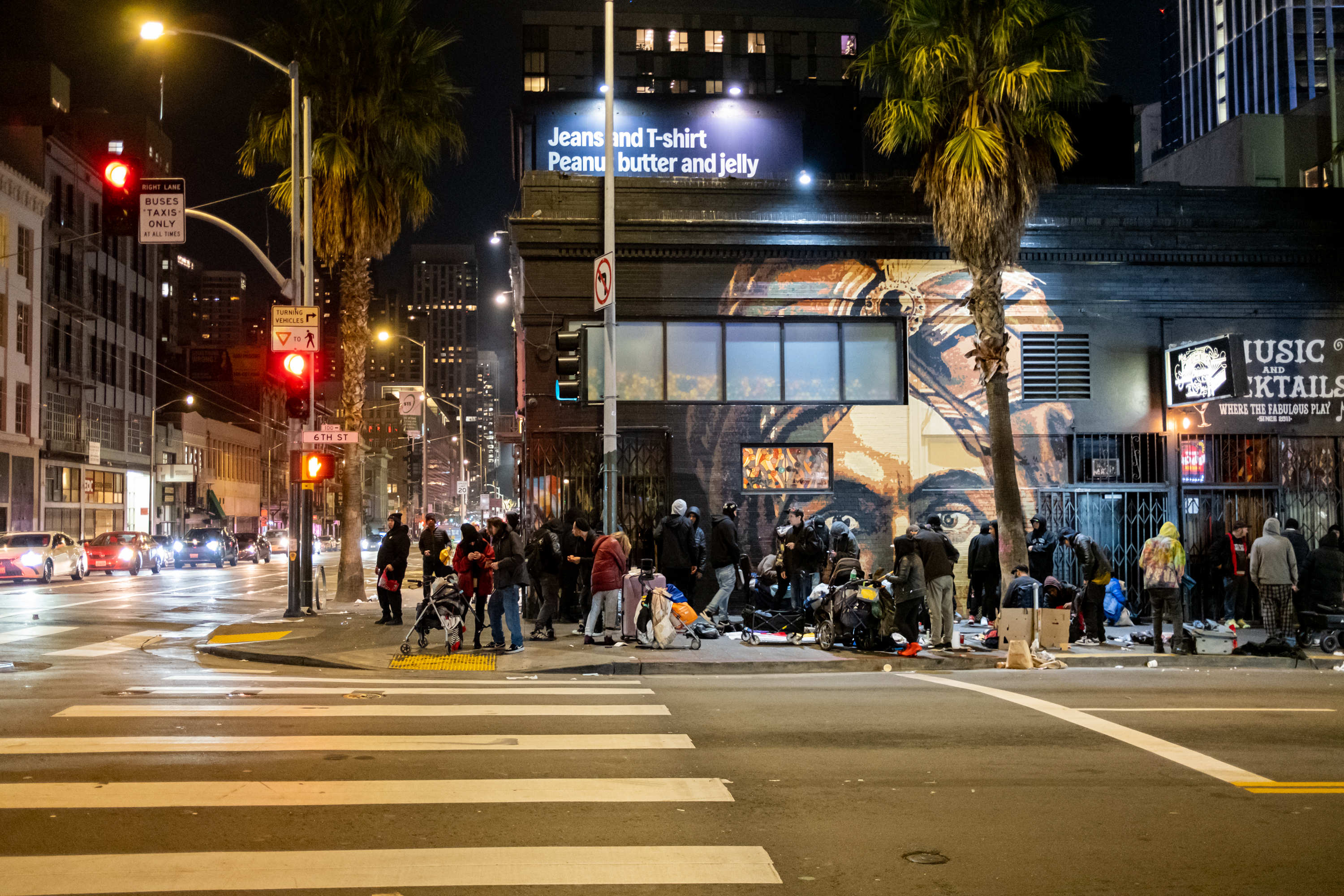 A nighttime city scene shows a group of people on a sidewalk next to a mural. They appear to be gathered around belongings, with traffic and buildings nearby.