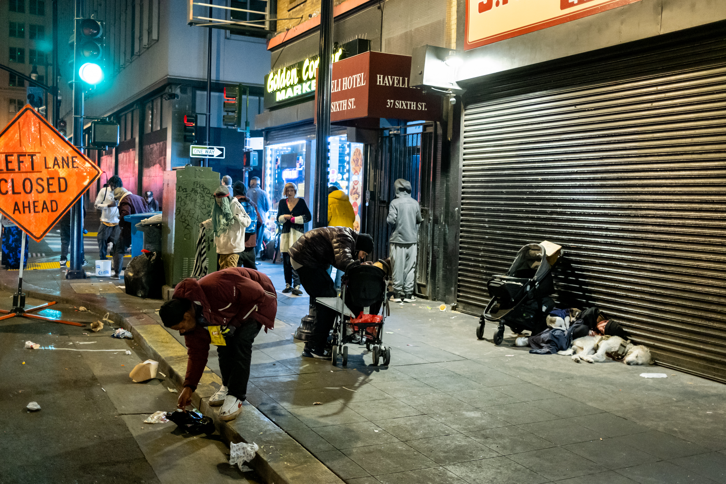 A nighttime street scene with people and strollers around, some bending down. A storefront with signs and a &quot;Left Lane Closed&quot; sign is visible.