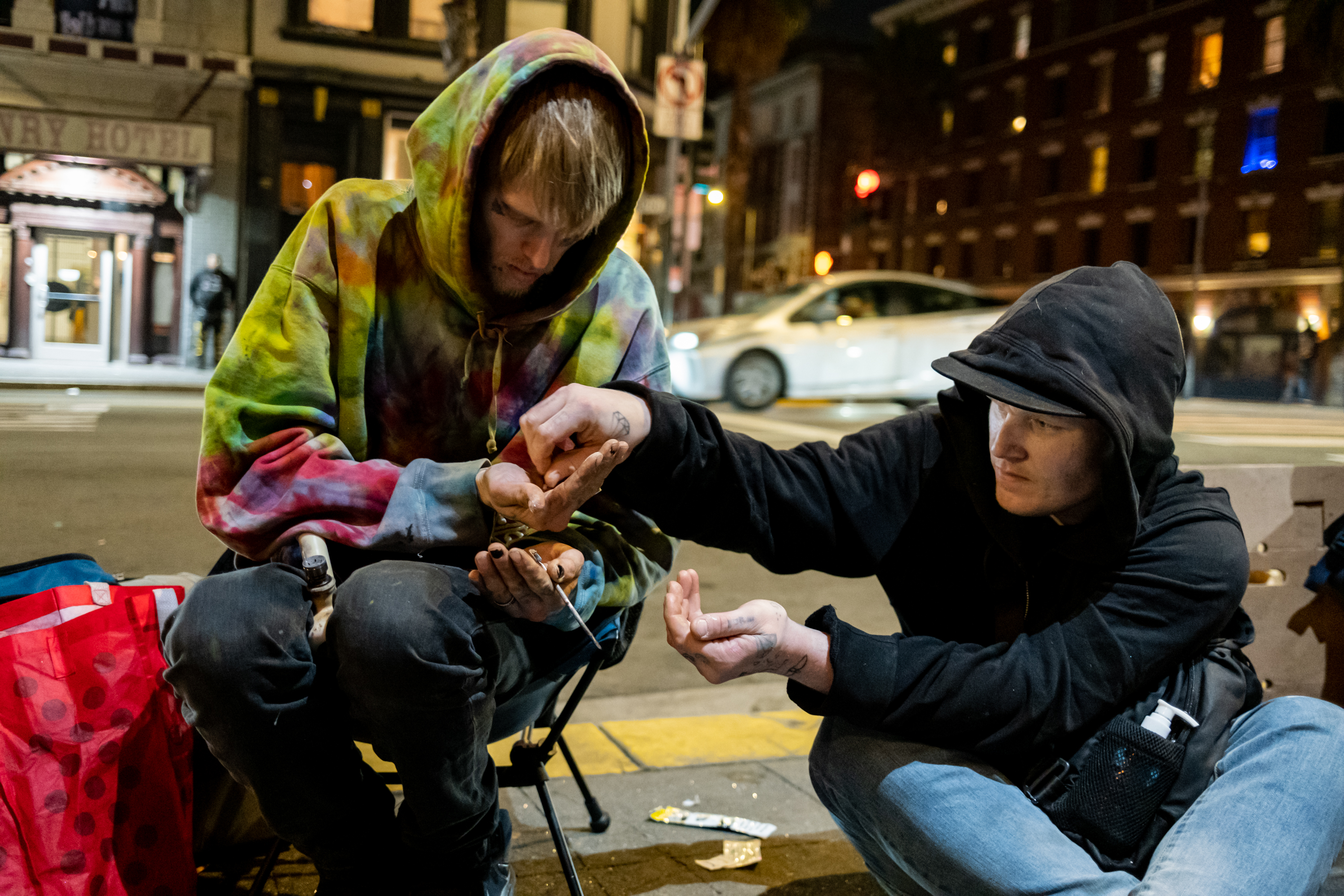 Two people sit on a sidewalk at night. One, in a tie-dye hoodie, holds something in their hand. The other, in a black hoodie, reaches out.