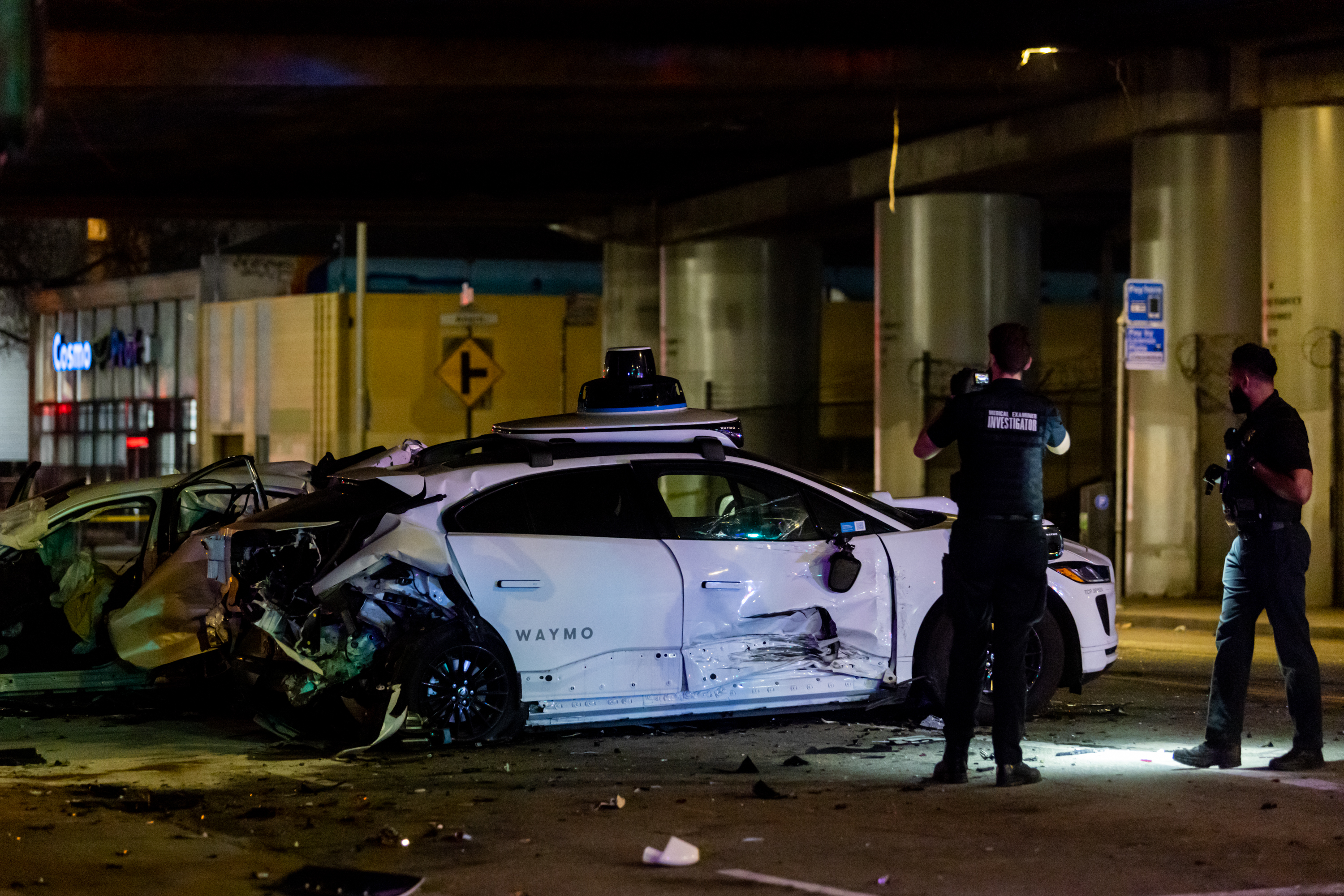 A damaged Waymo car is involved in a collision under a bridge, surrounded by debris. Two investigators in dark clothing examine and photograph the scene.