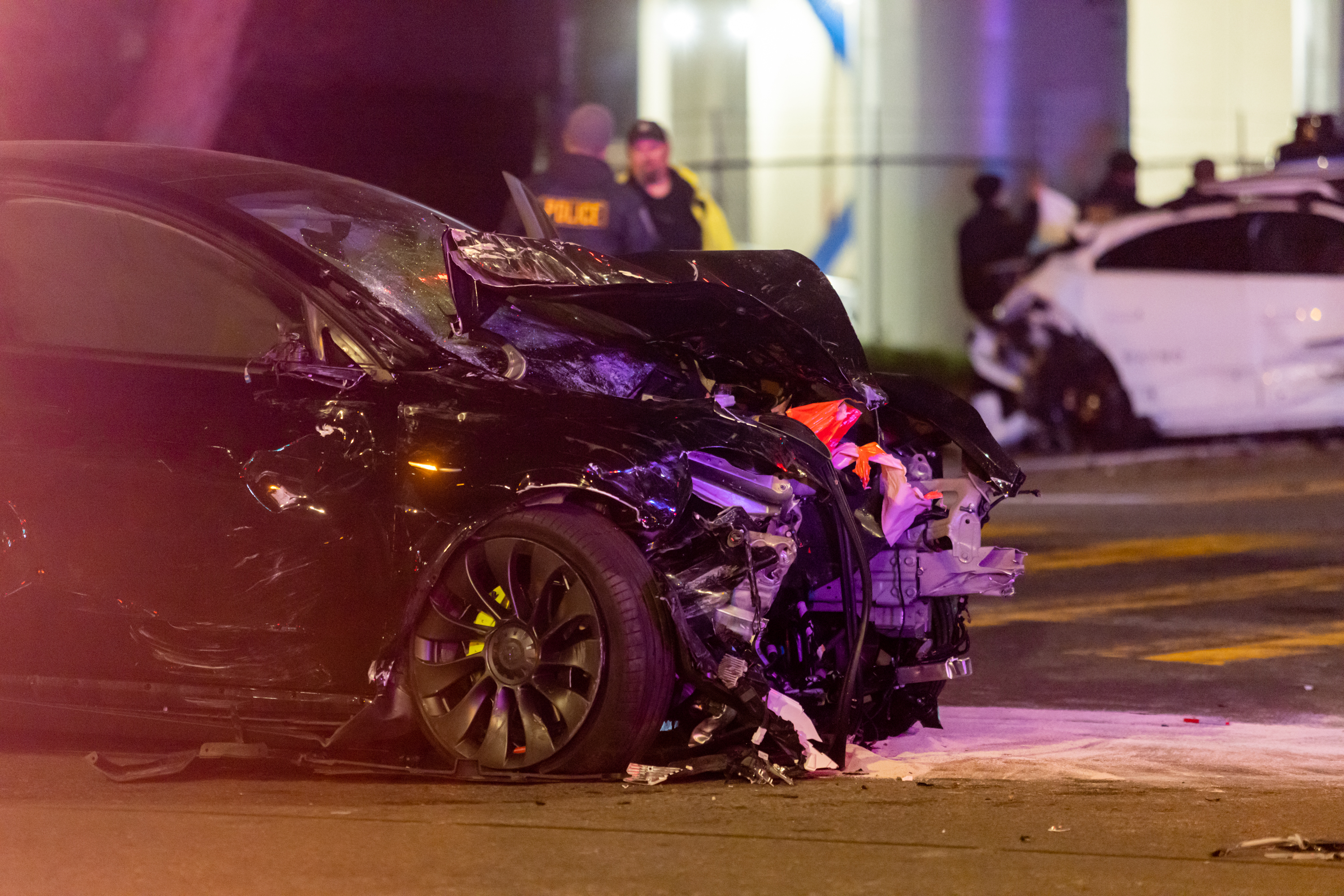 A black car with severe front damage is shown at night, with police officers in the background and another damaged white car visible further back.