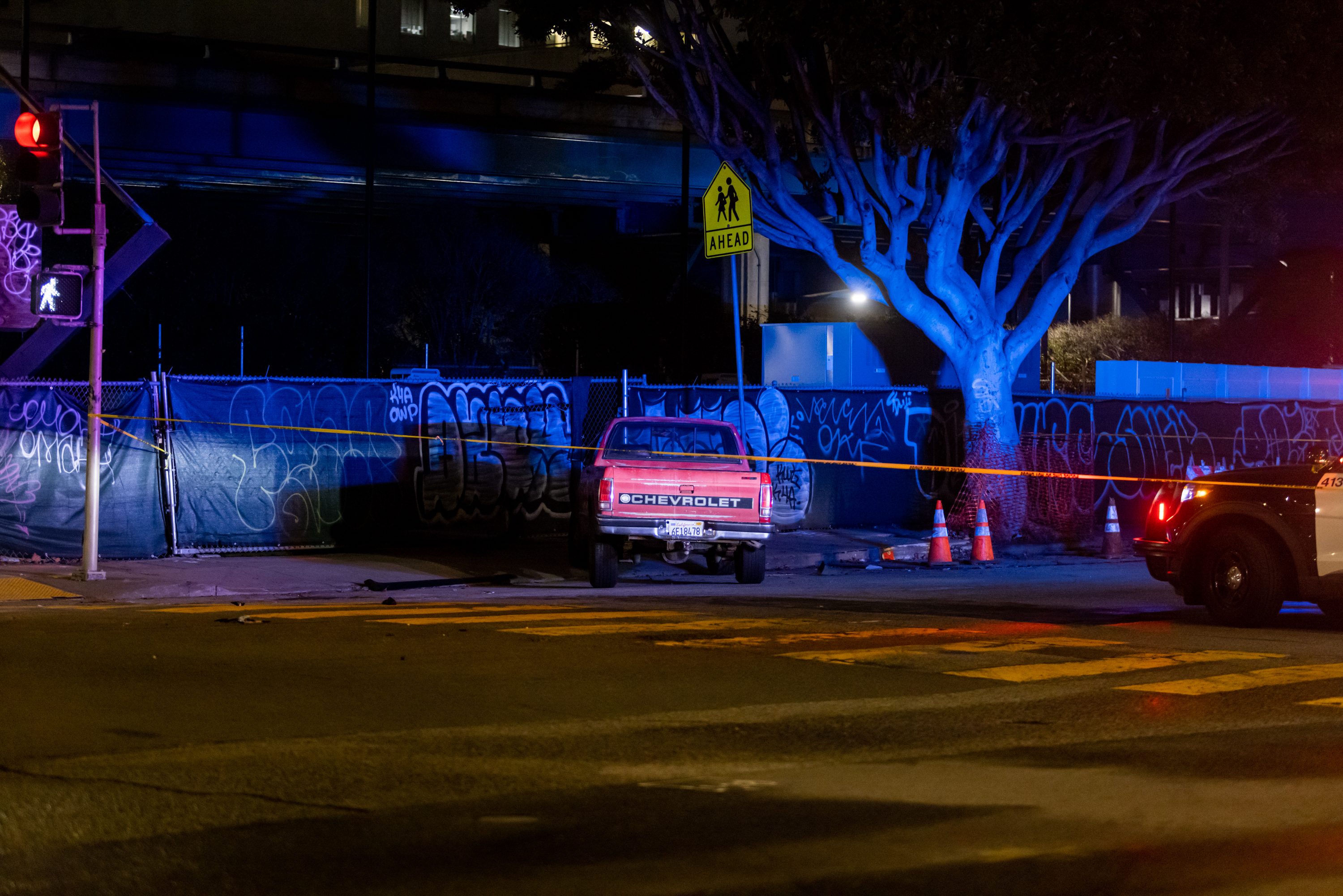 A red Chevrolet truck is parked by a graffiti-covered fence. A police car with flashing lights is nearby, and caution tape surrounds the scene at night.