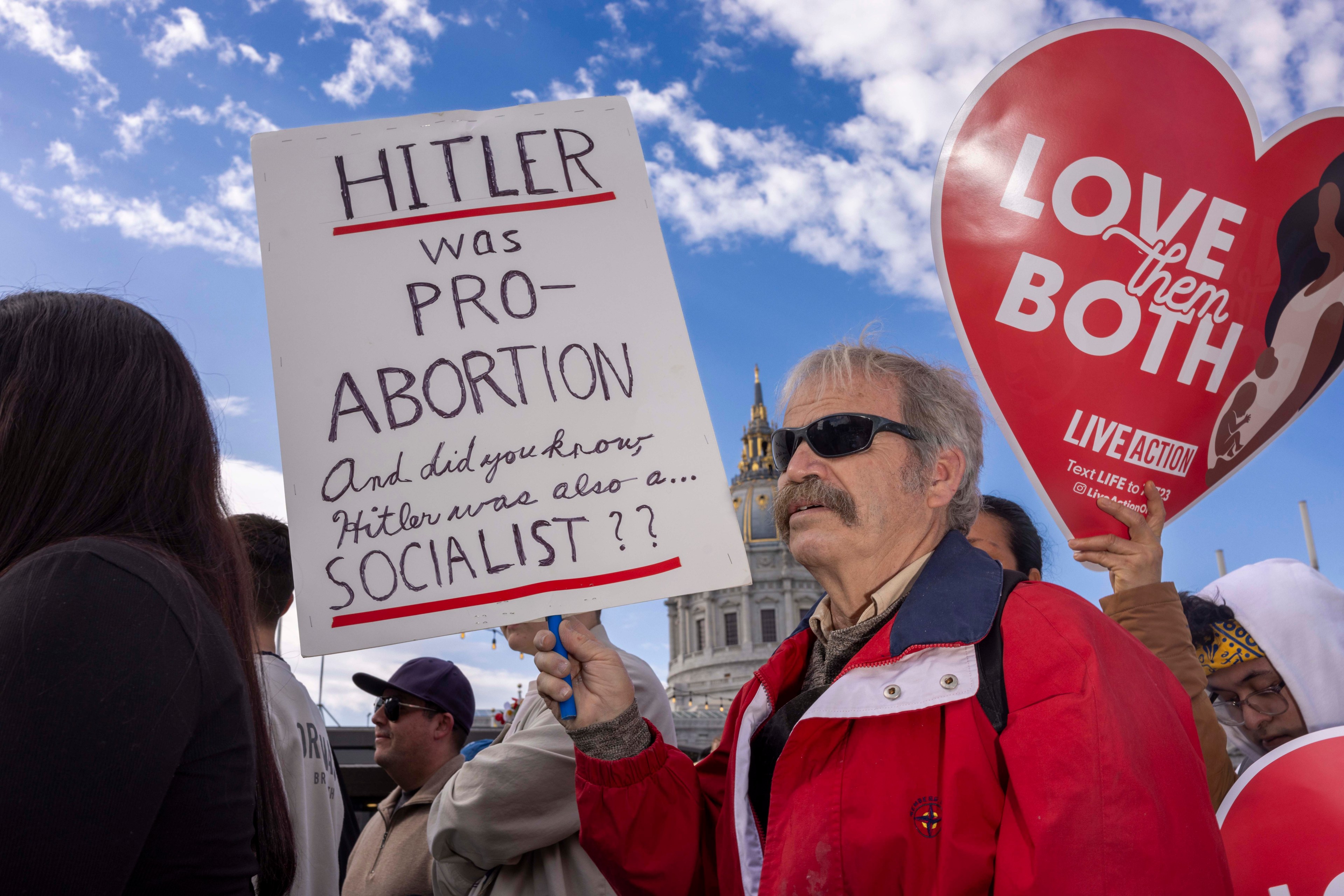 A man in a red jacket holds a sign about Hitler and socialism during a protest. Another person holds a heart-shaped sign saying &quot;Love them both,&quot; against a clear sky.