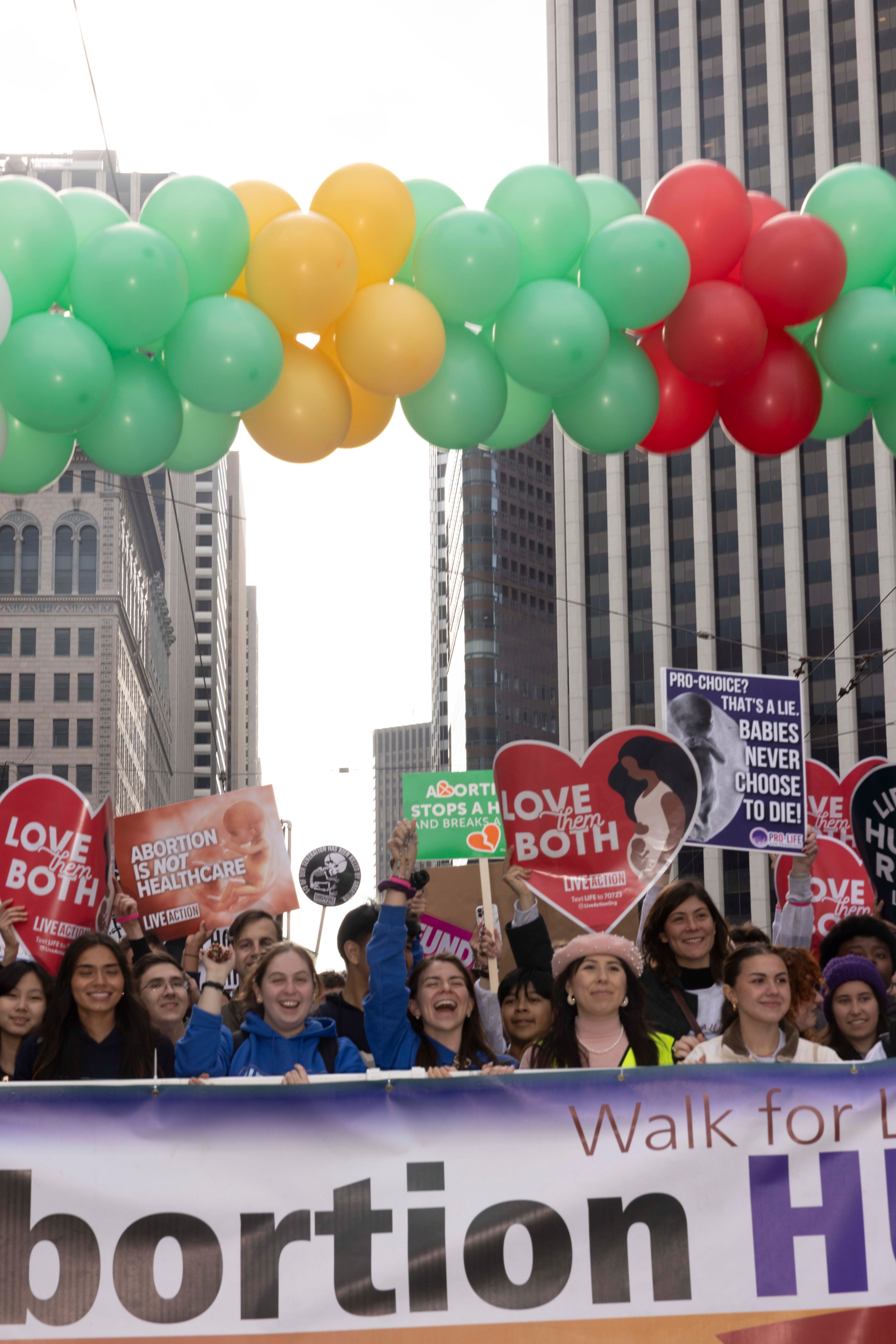 A group of people in an urban setting hold signs with anti-abortion messages. Colorful balloons form an arch overhead, and buildings are visible in the background.