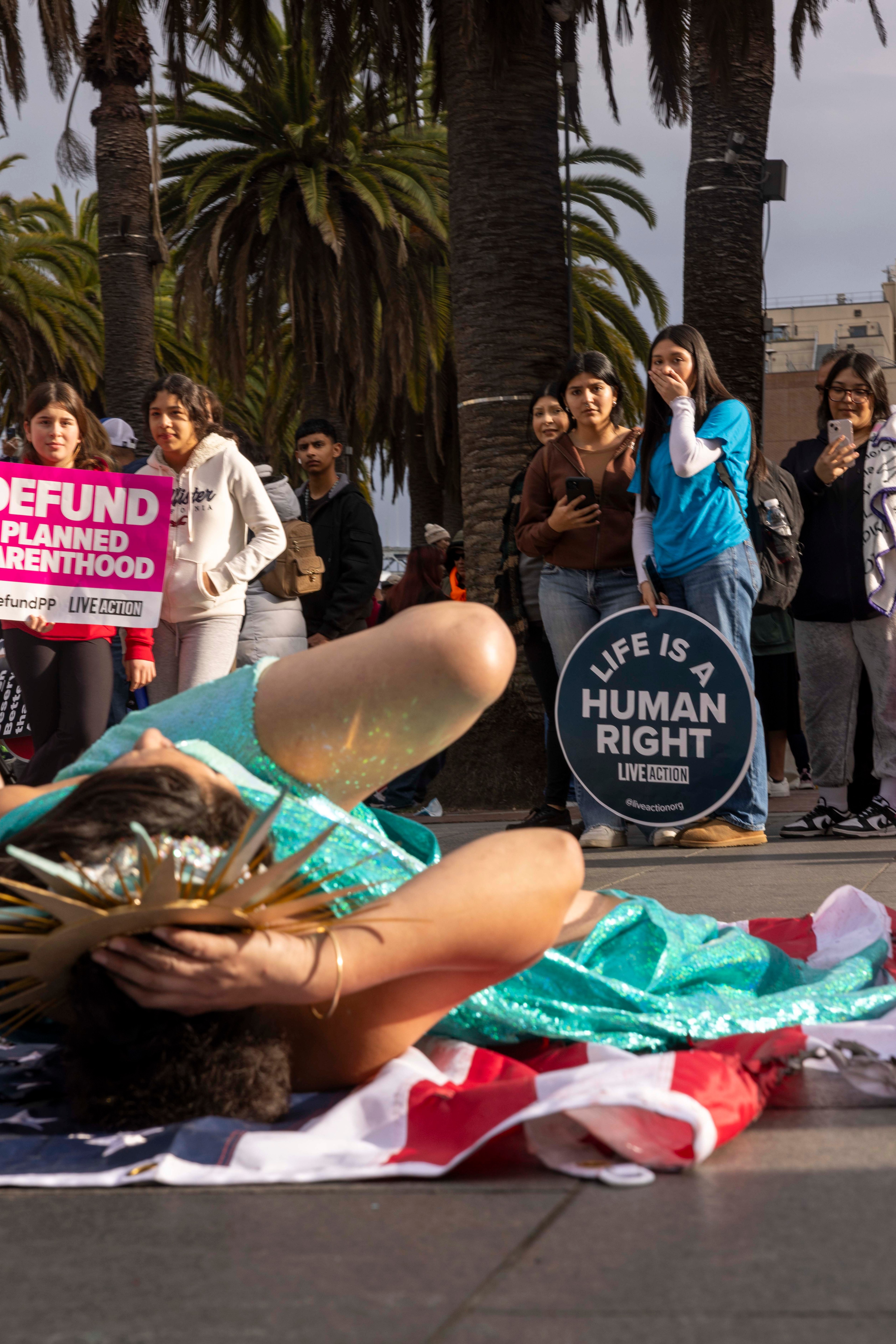 A person in a teal costume lies on an American flag, surrounded by people holding signs that read &quot;Defund Planned Parenthood&quot; and &quot;Life is a Human Right.&quot;