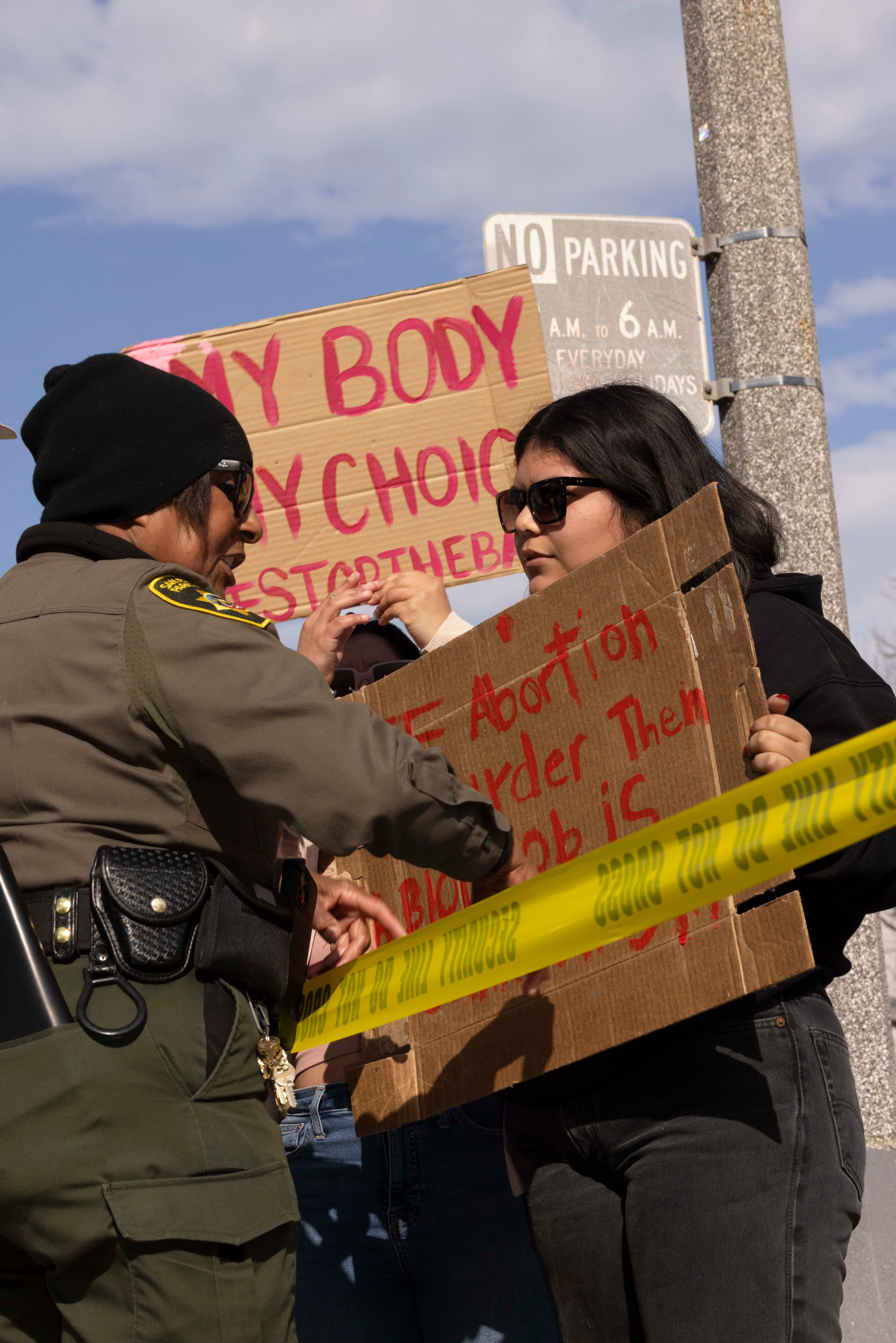 A police officer and a protester holding signs engage behind yellow tape. The signs display messages about body autonomy and abortion rights.