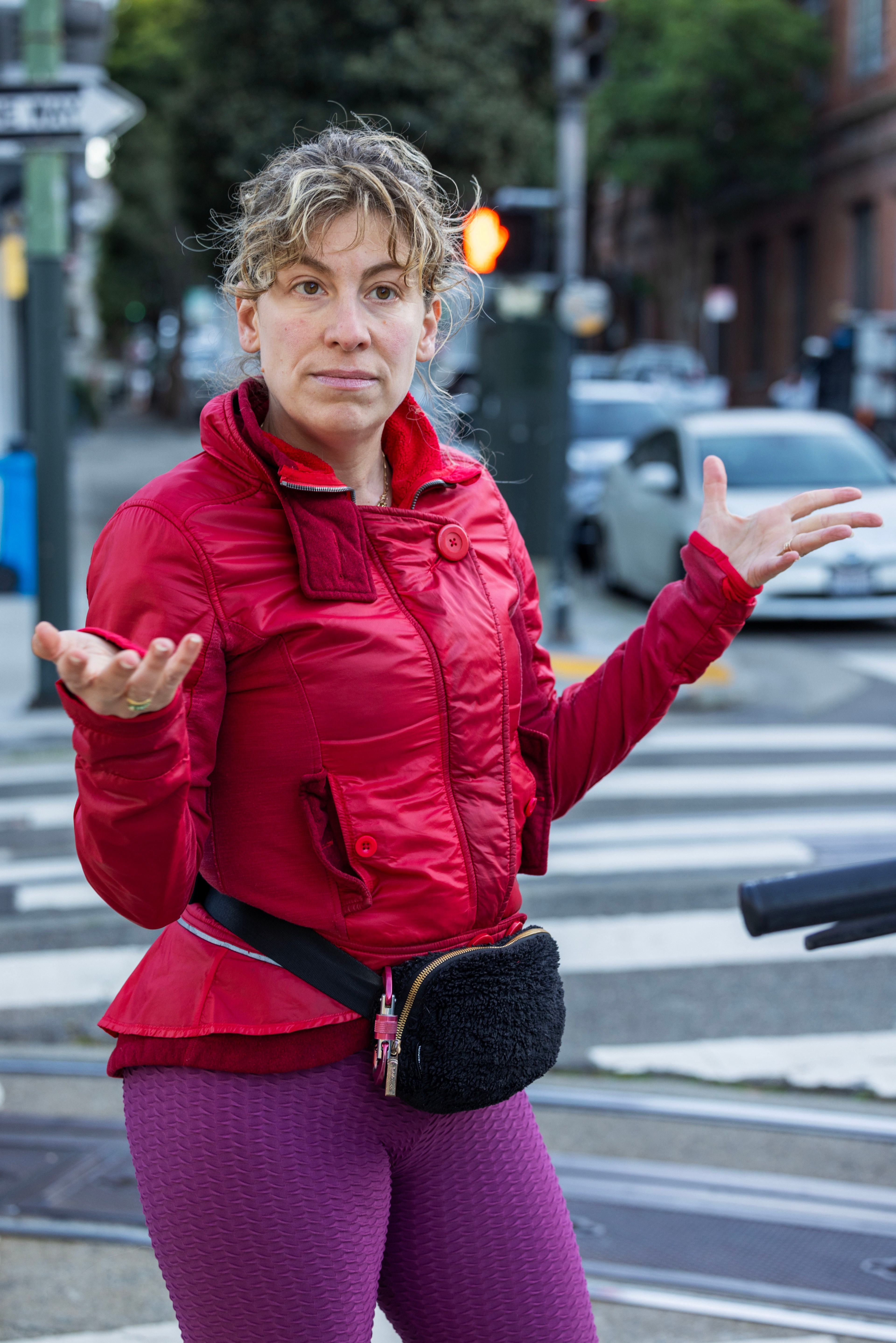 A woman in a red jacket stands at a street crossing, gesturing with her hands. She has curly hair, wears purple leggings, and has a black fanny pack.