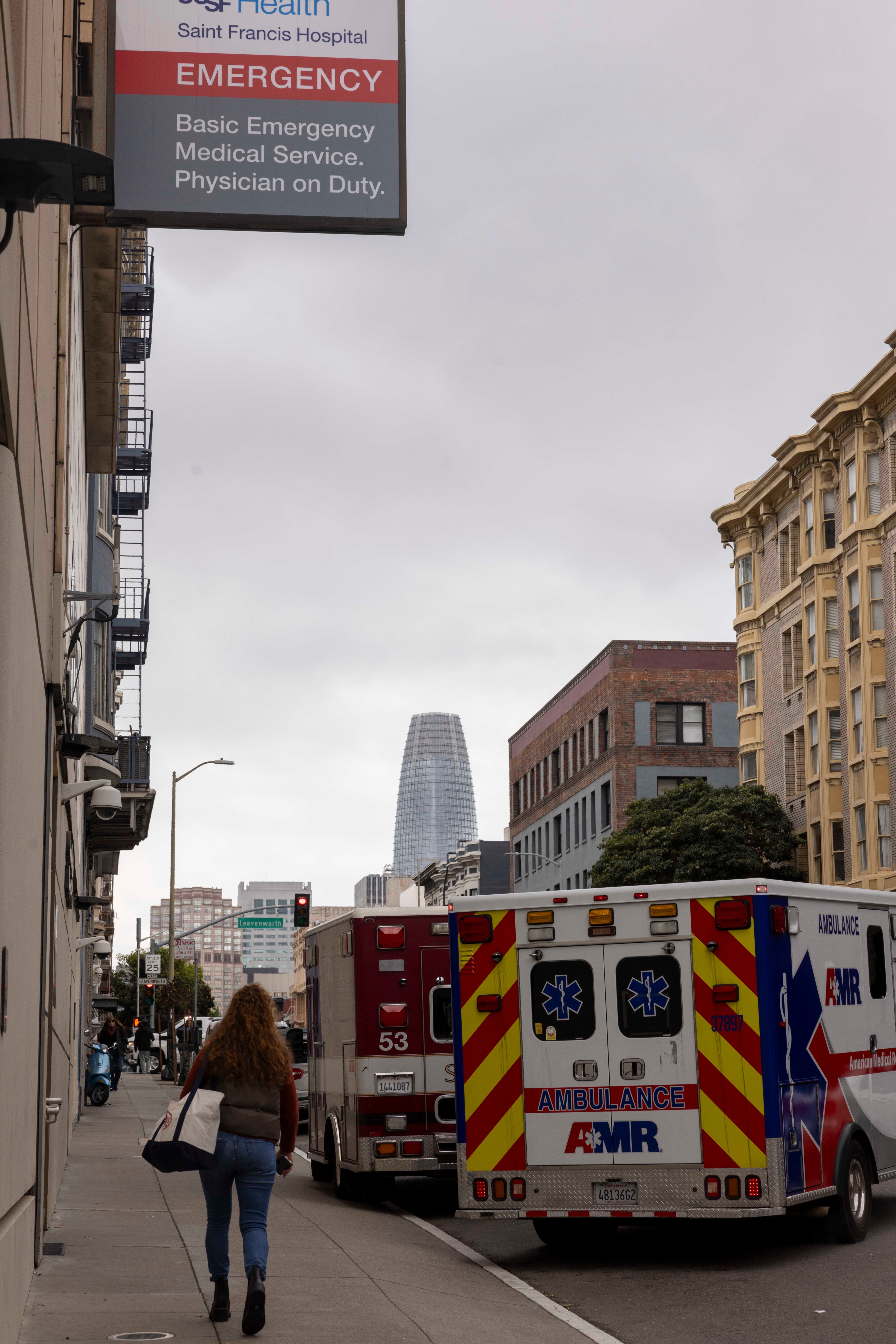 A woman walks along a city sidewalk with ambulances parked nearby. A hospital sign reads &quot;Emergency&quot; and a tall, modern building looms in the background.