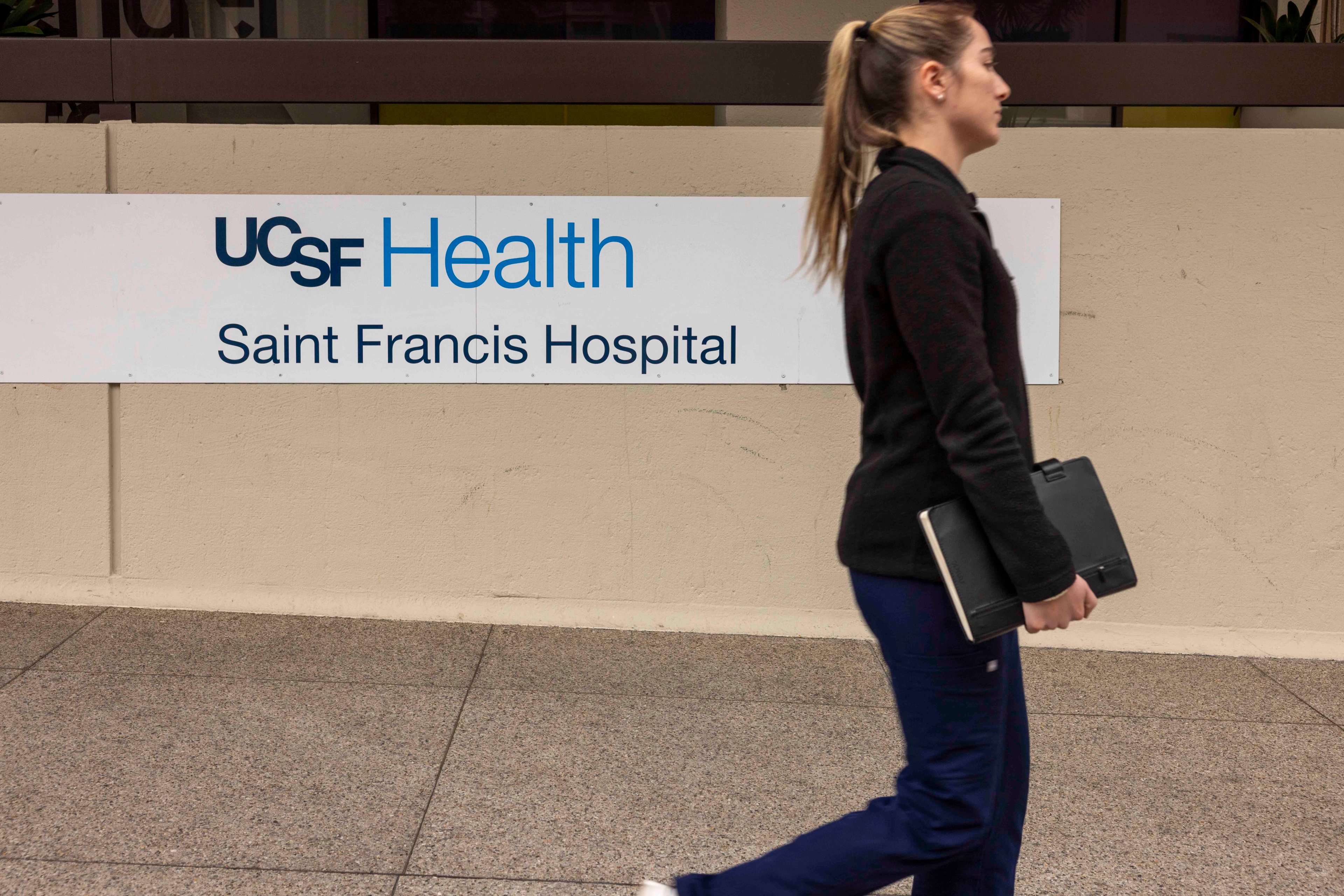 A woman is walking past a sign for UCSF Health Saint Francis Hospital, holding a closed laptop against her side.