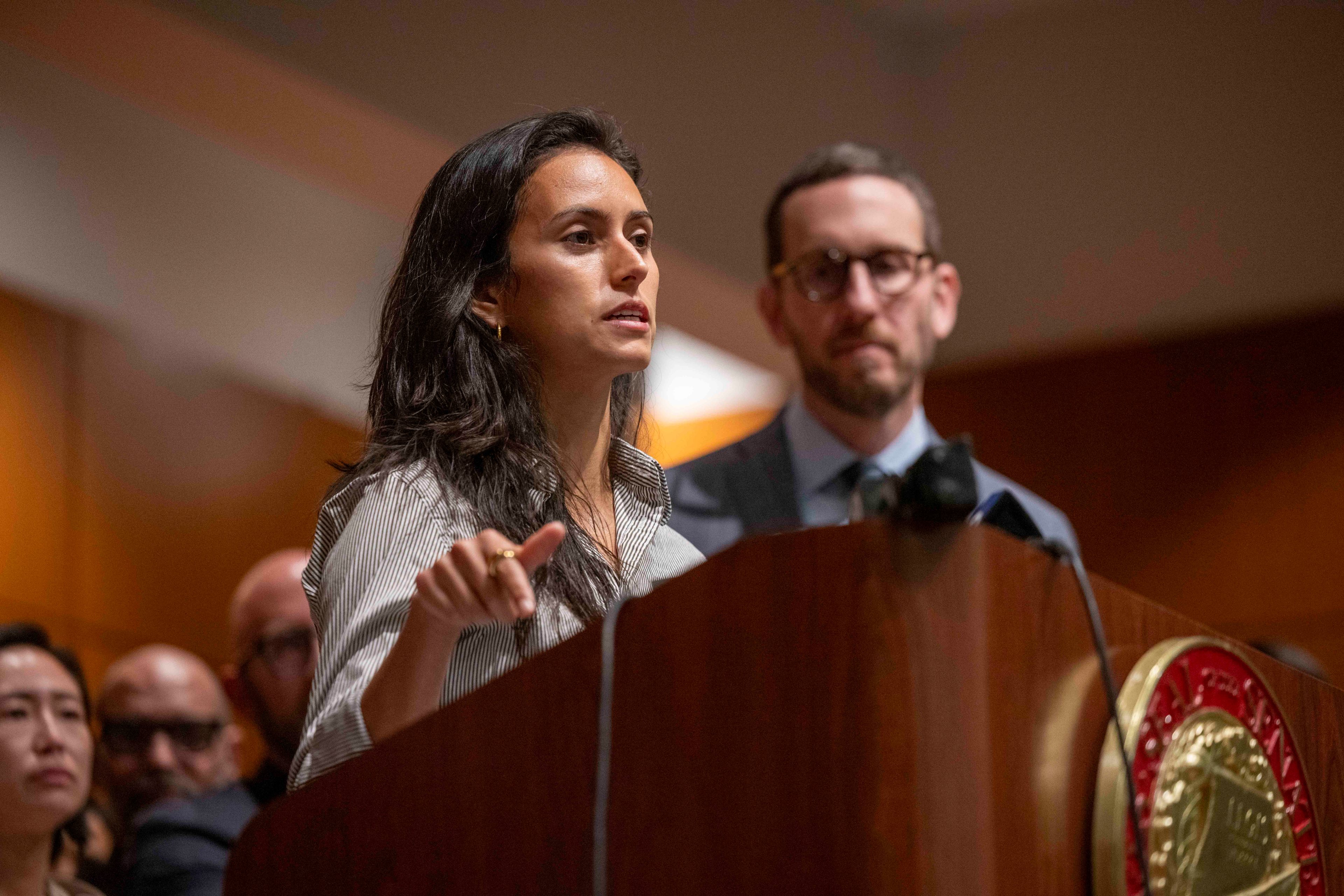 A woman speaks passionately at a podium, pointing, with a man standing behind her. The scene appears to be a formal event, with several people in the background.