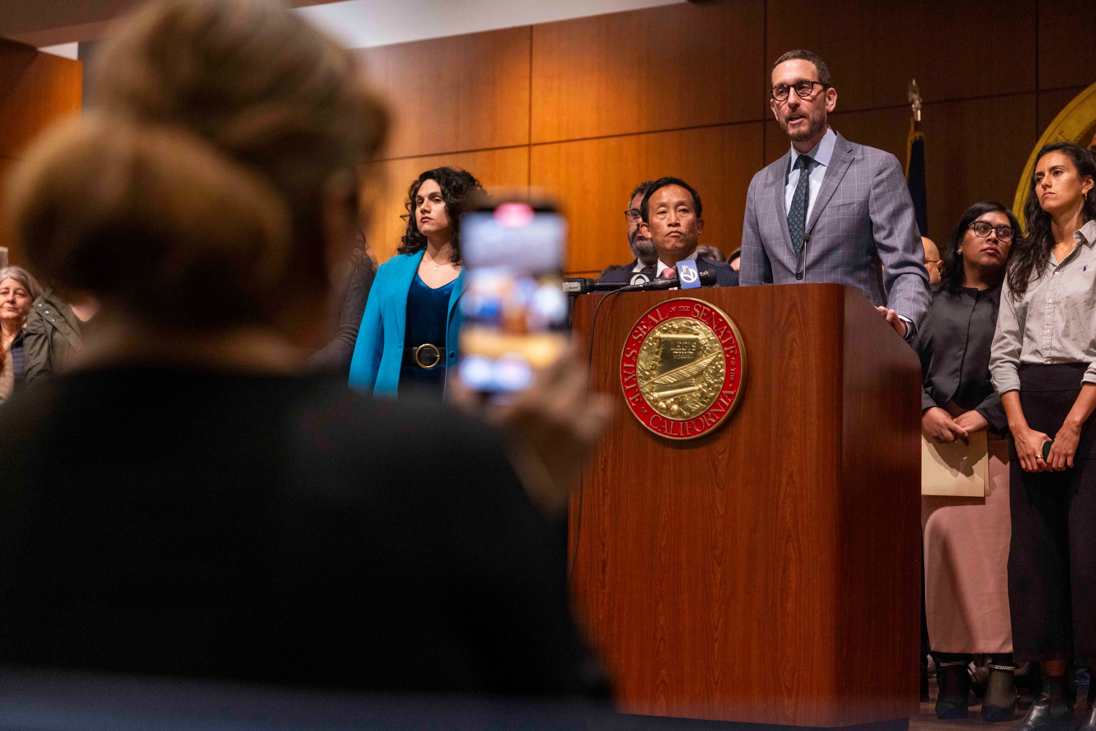 A group of people stand at a podium with microphones in a formal setting. A person in the foreground is taking a photo with a smartphone.