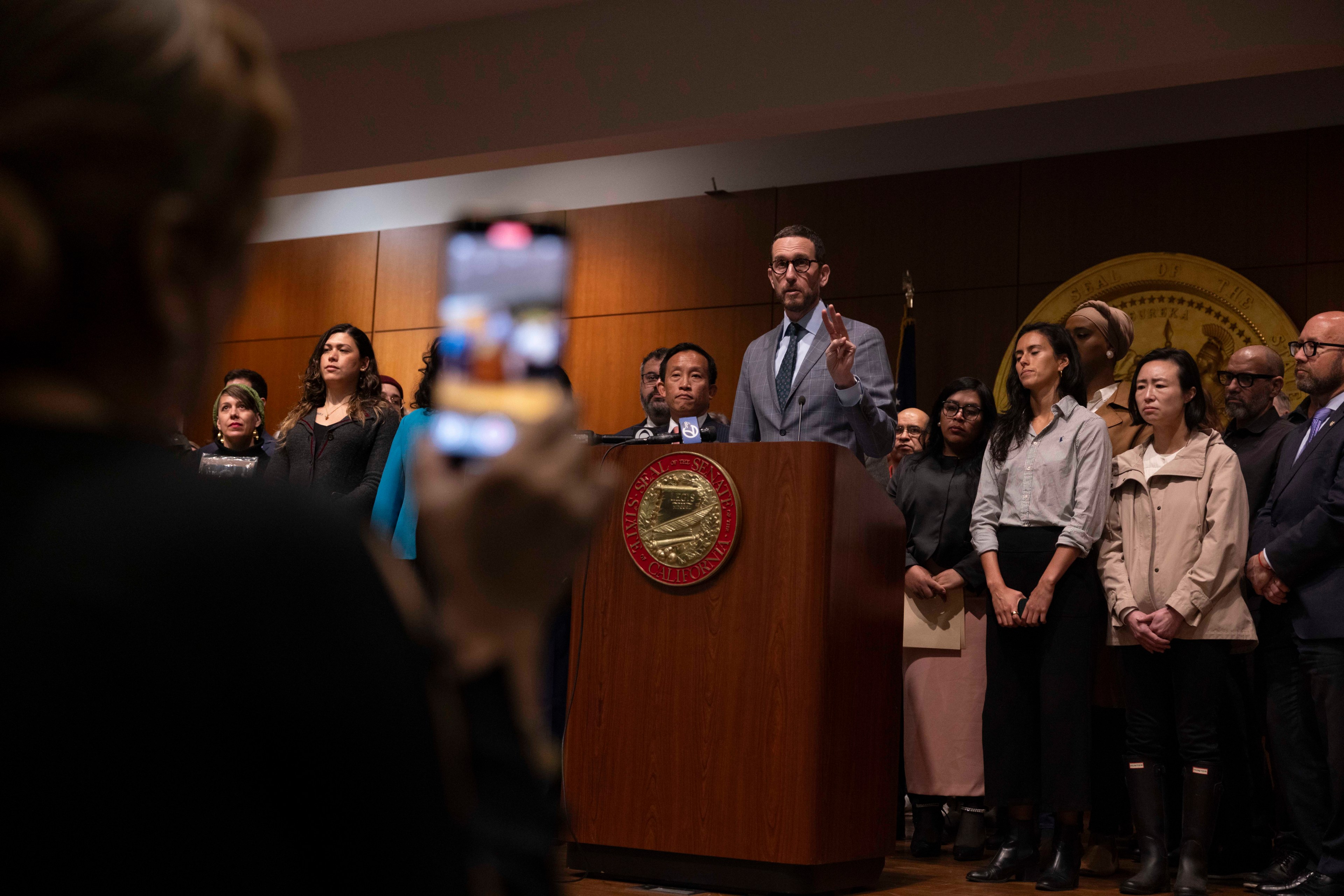 A tall man in a gray suit delivers remarks at a wooden podium flanked by a crowd of people.