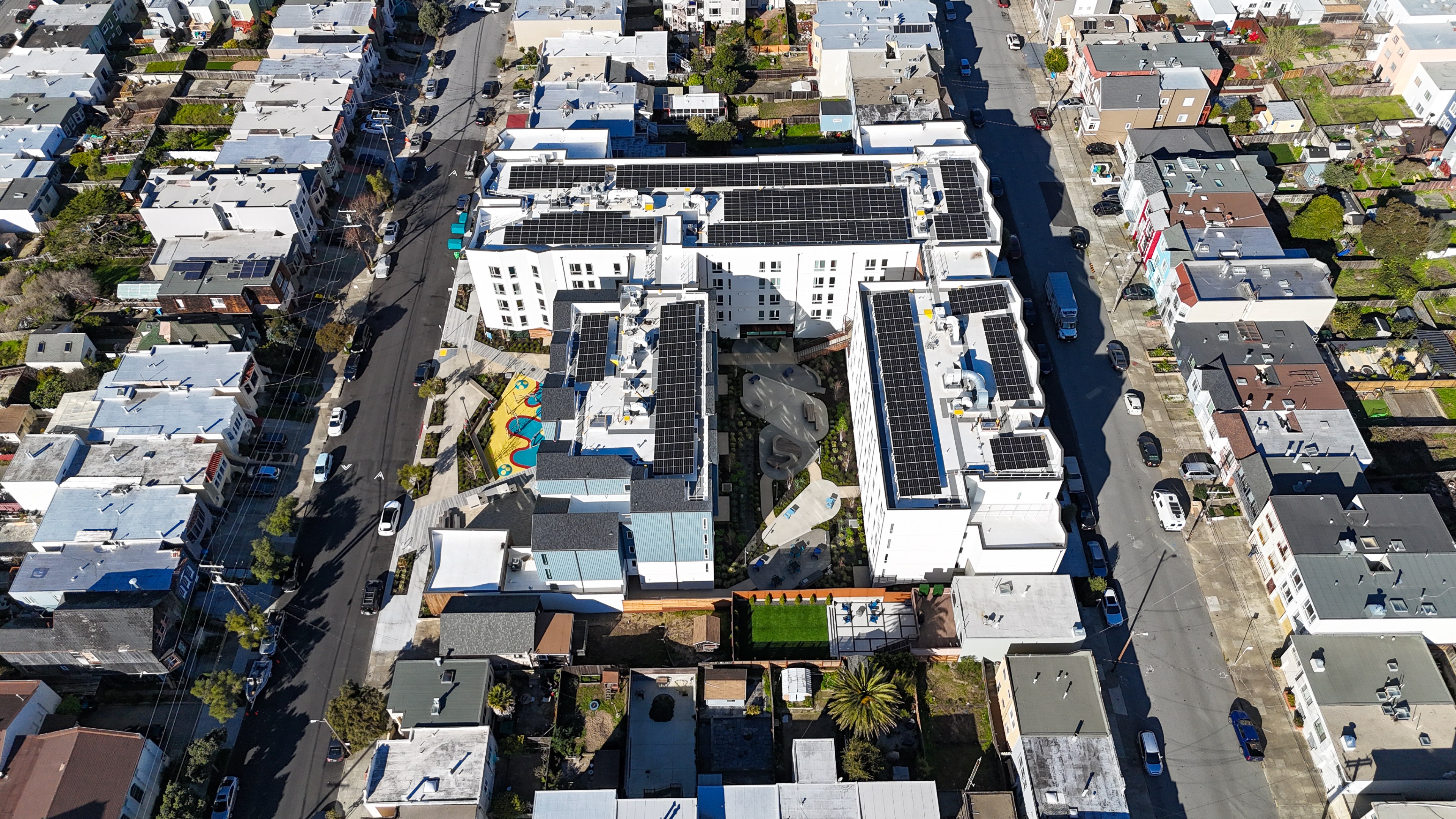 The image shows an aerial view of a residential area with white buildings, some sporting solar panels. A colorful playground and greenery are visible between structures.