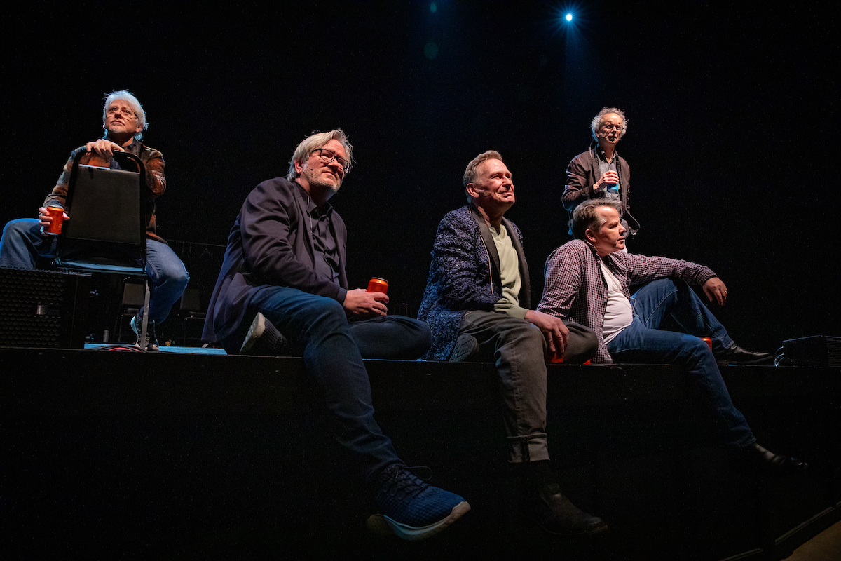 Five men are casually seated on a stage, some holding drinks. They appear relaxed and engaged, with dim lighting above creating a cozy atmosphere.