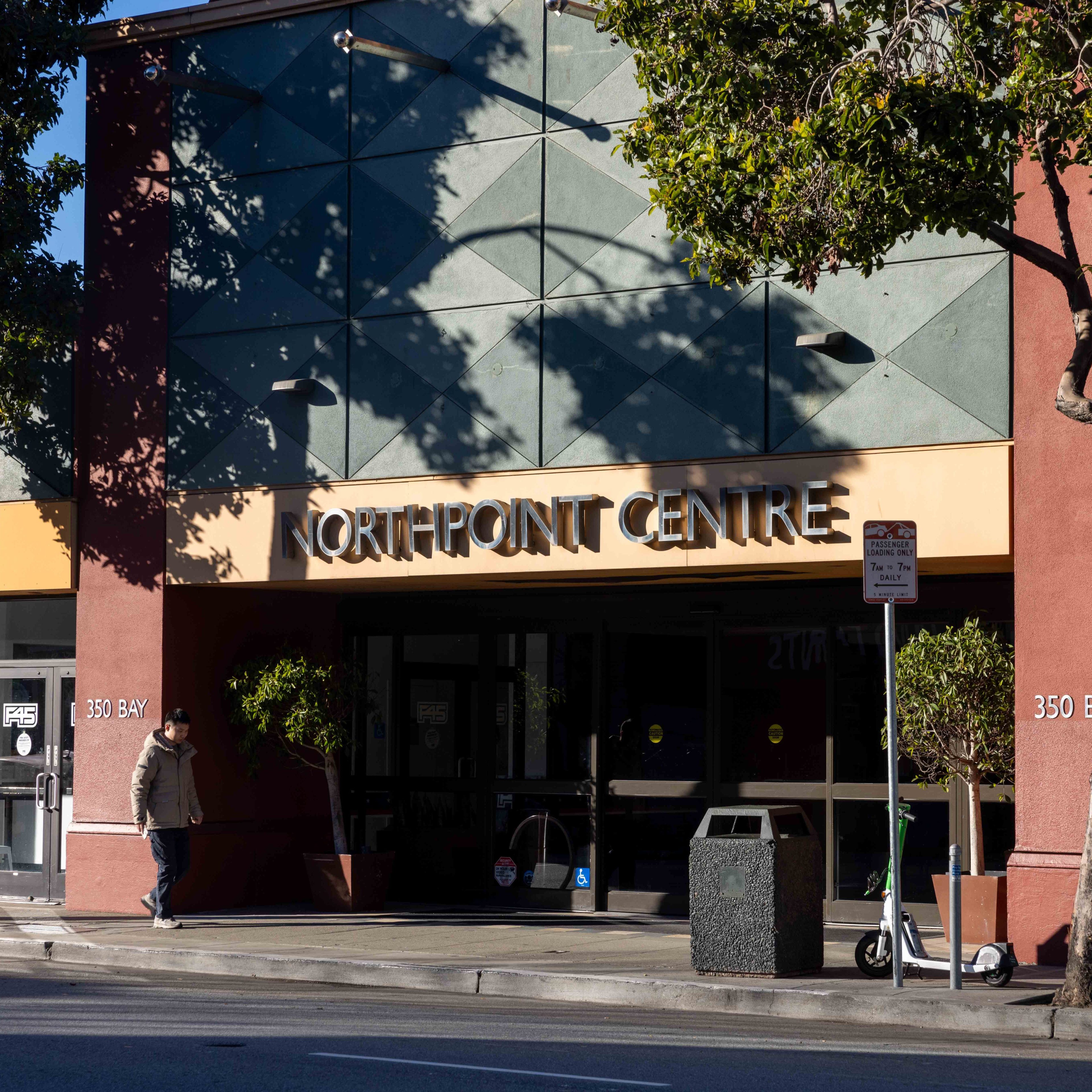 A man walks past the Northpoint Centre, a building with geometric designs and a shaded entrance. A scooter and trash can are nearby, along with a street sign.