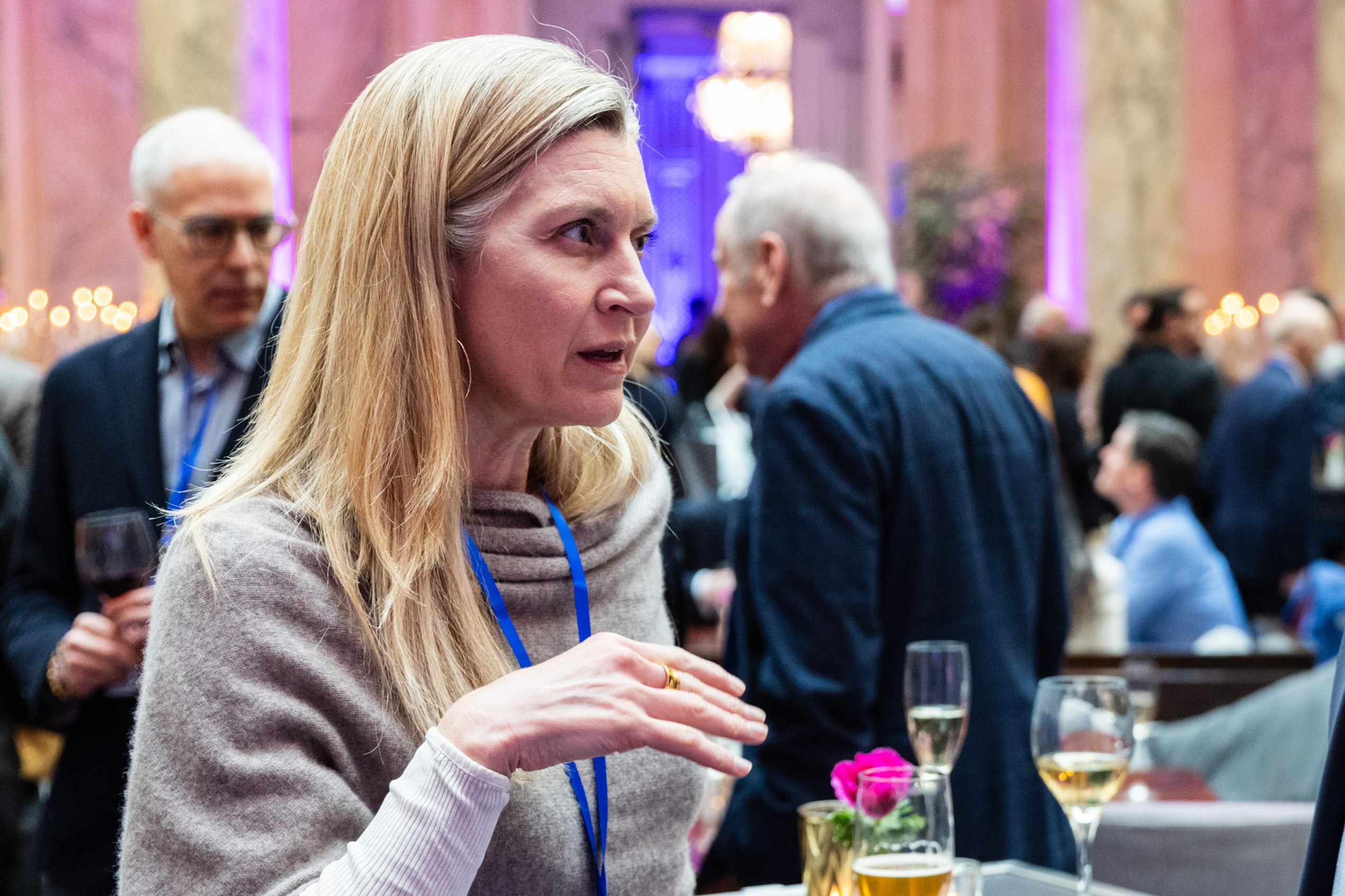 A woman is engaged in conversation at an event. She wears a light sweater and has a blue lanyard. People in the background are holding drinks, with dim, colorful lighting.