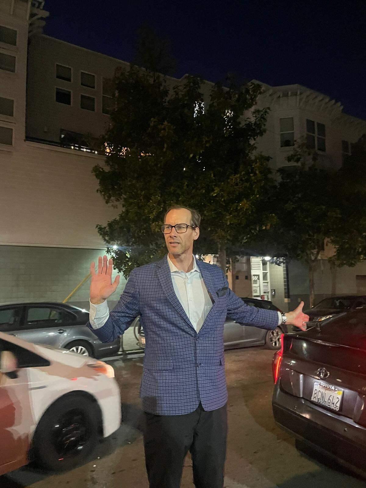 A man in a blue blazer stands on a city street at night with arms outstretched; cars are parked nearby, and an apartment building is in the background.