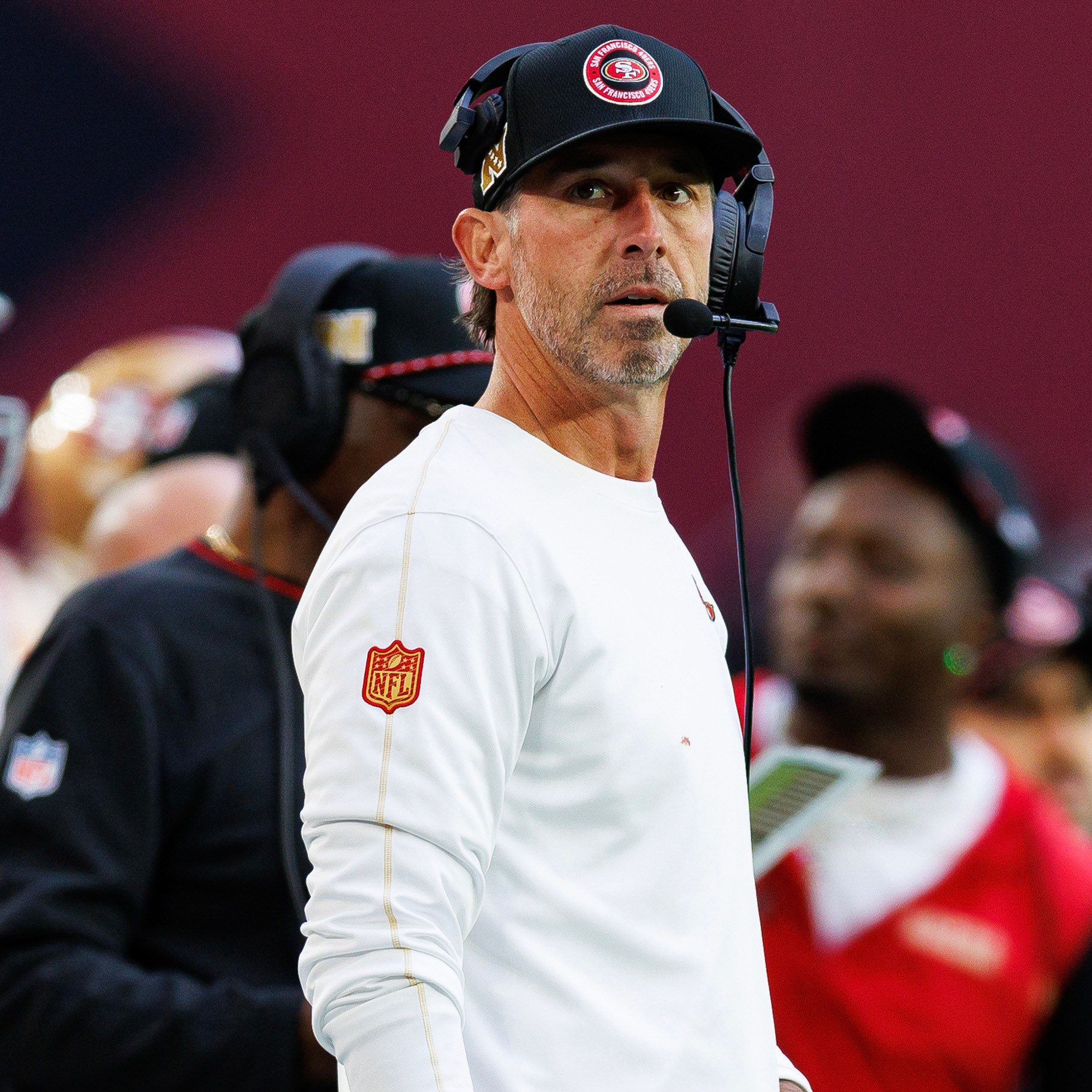 A focused football coach wearing a headset and a team cap stands on the sidelines, with players and staff in the background during a game.