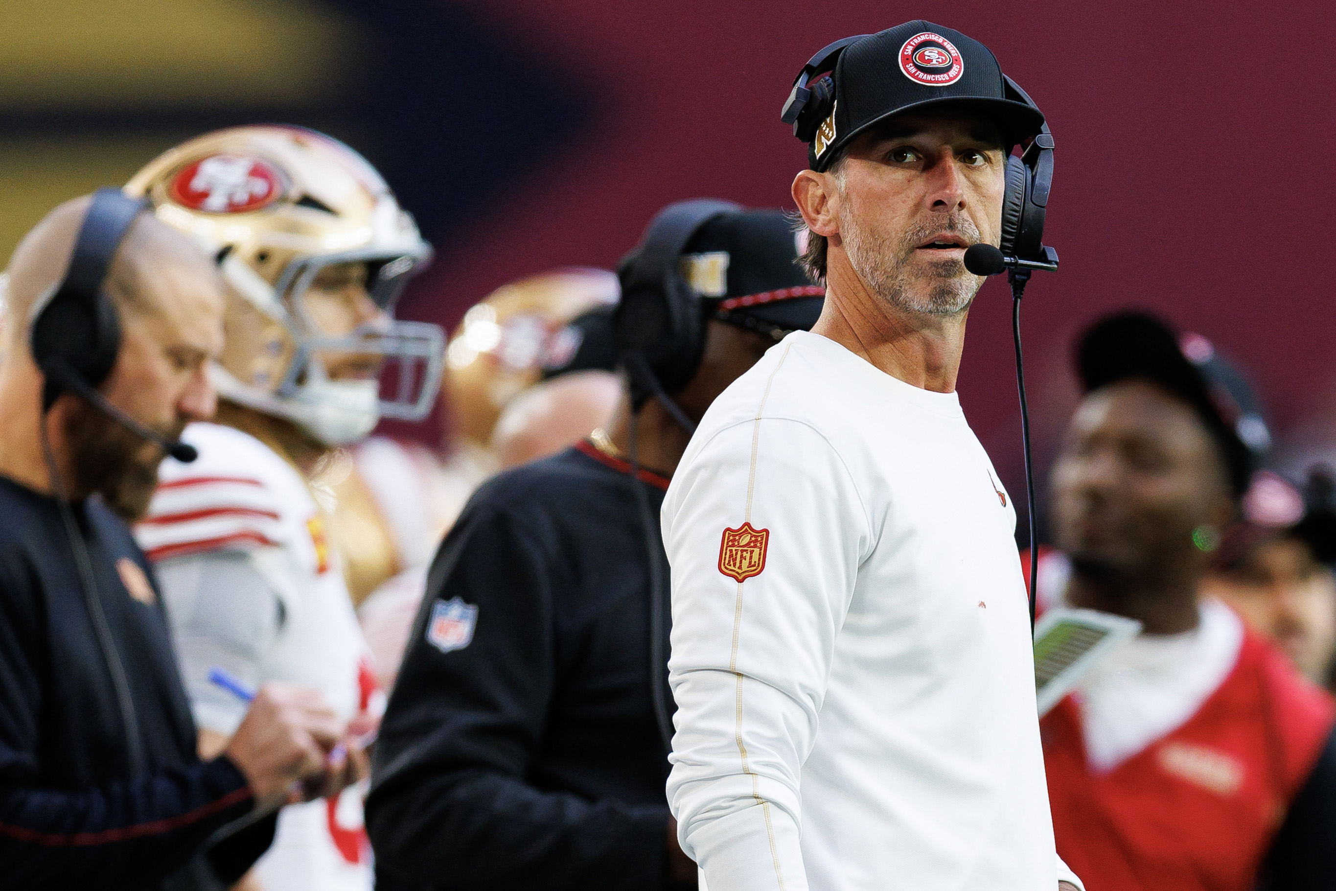 A focused football coach wearing a headset and a team cap stands on the sidelines, with players and staff in the background during a game.