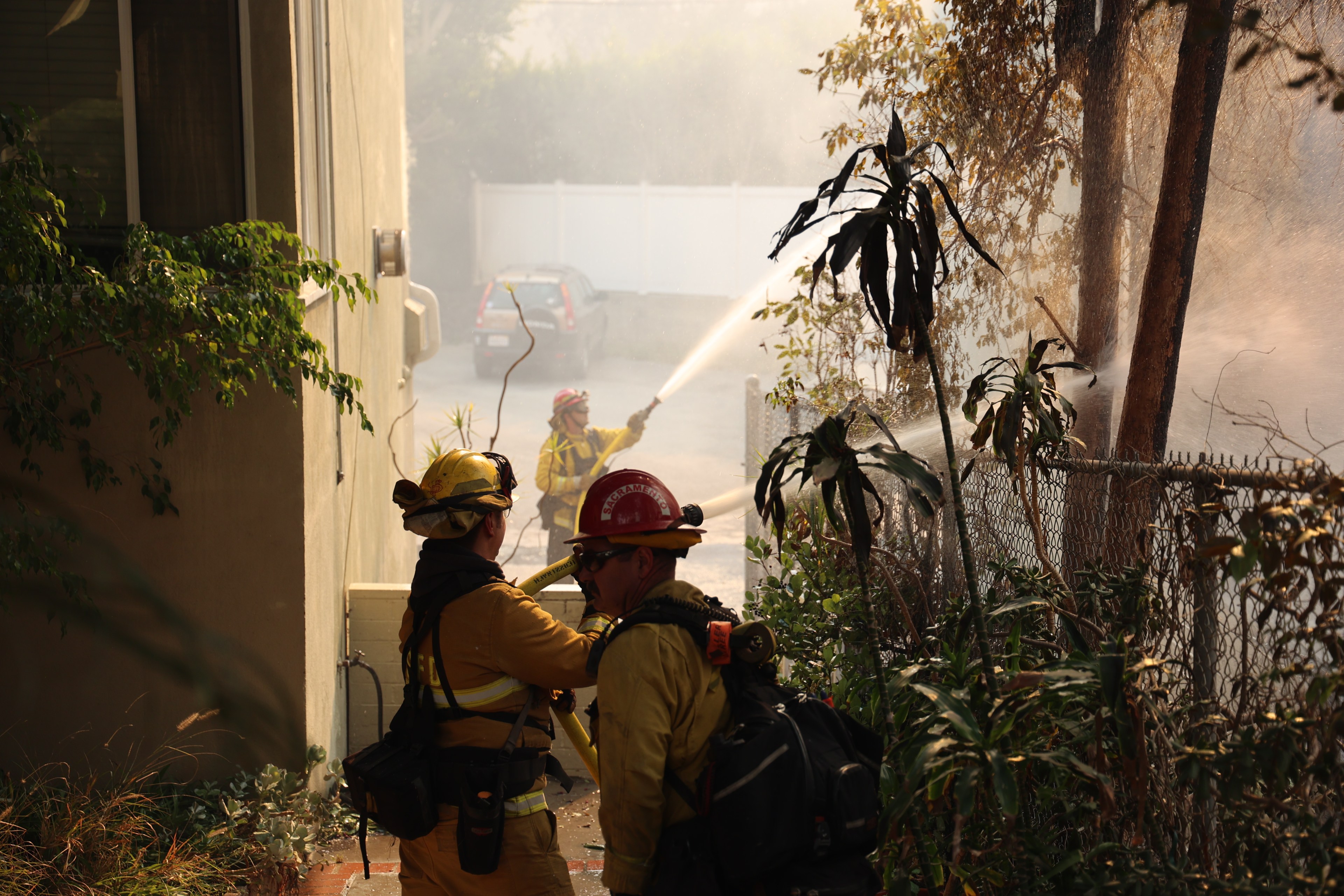 Firefighters in yellow gear spray water on trees near a building. Smoke fills the air and a gray car is visible in the background.