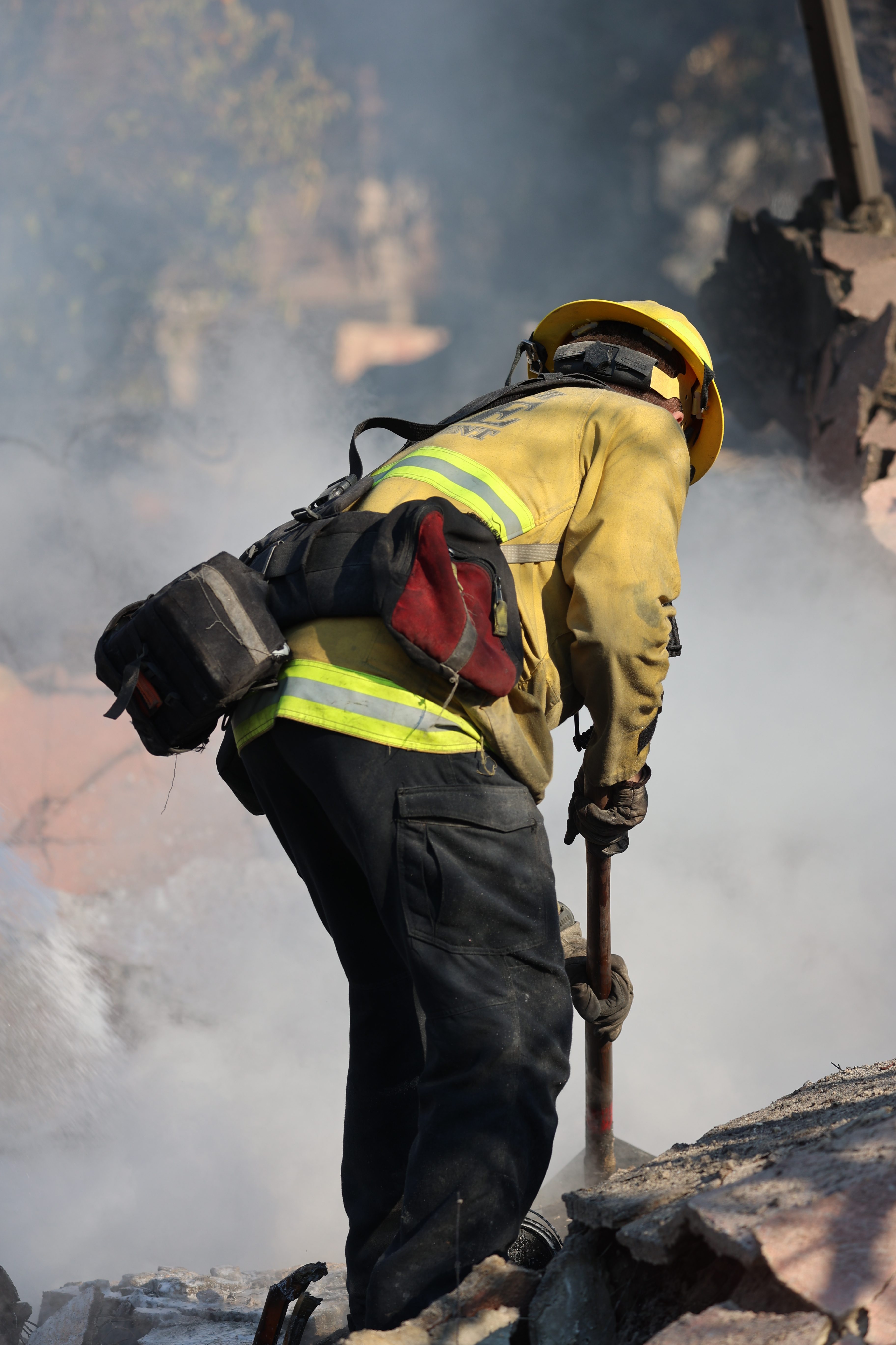 A firefighter in a yellow helmet and jacket stands amidst smoke, using a tool on rubble.